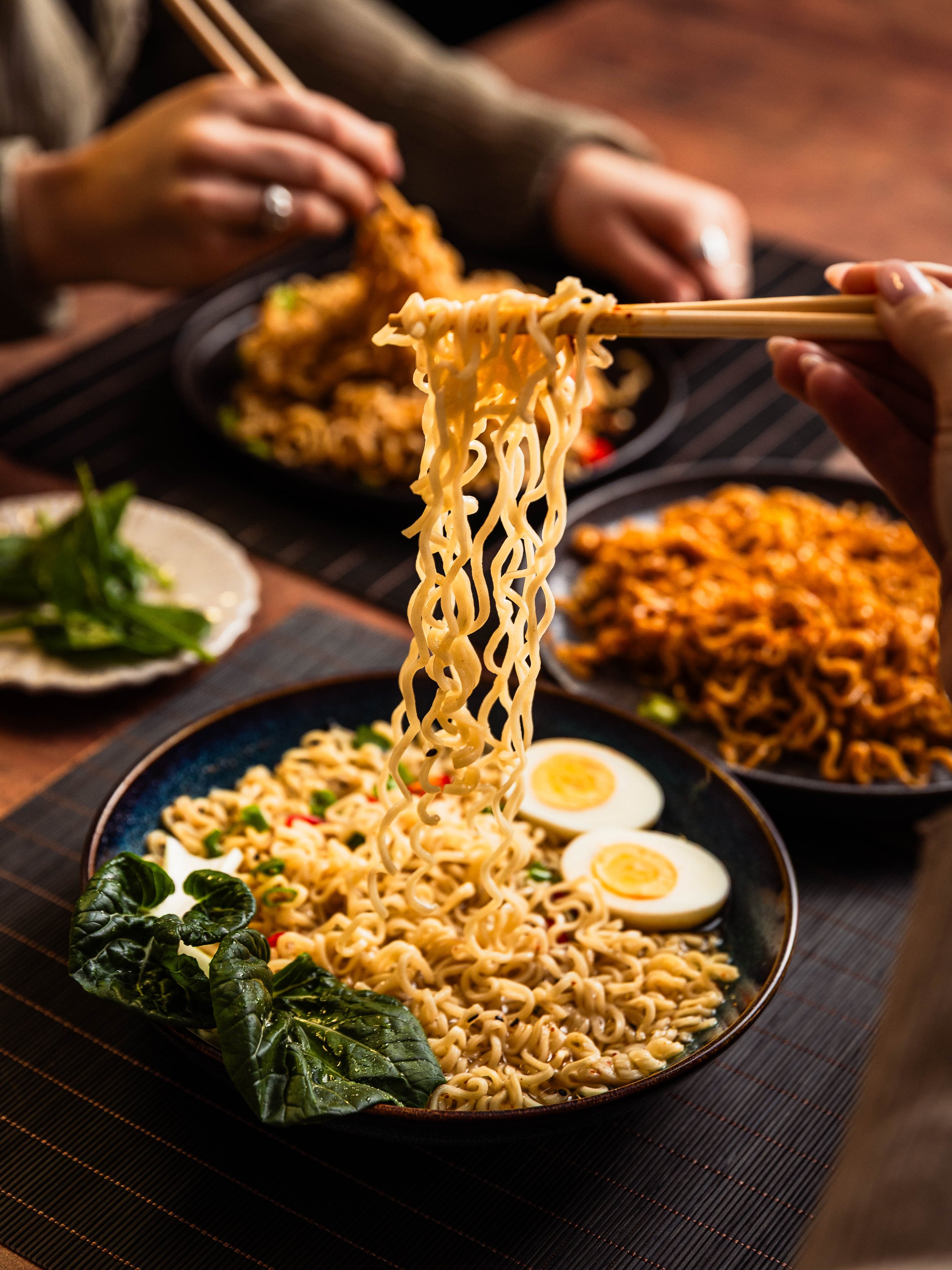 A bowl of ramen noodles with boiled eggs, leafy greens, and chopped vegetables on a dark placemat, with hands using chopsticks to lift noodles, and additional dishes of fried rice and other foods in the background.