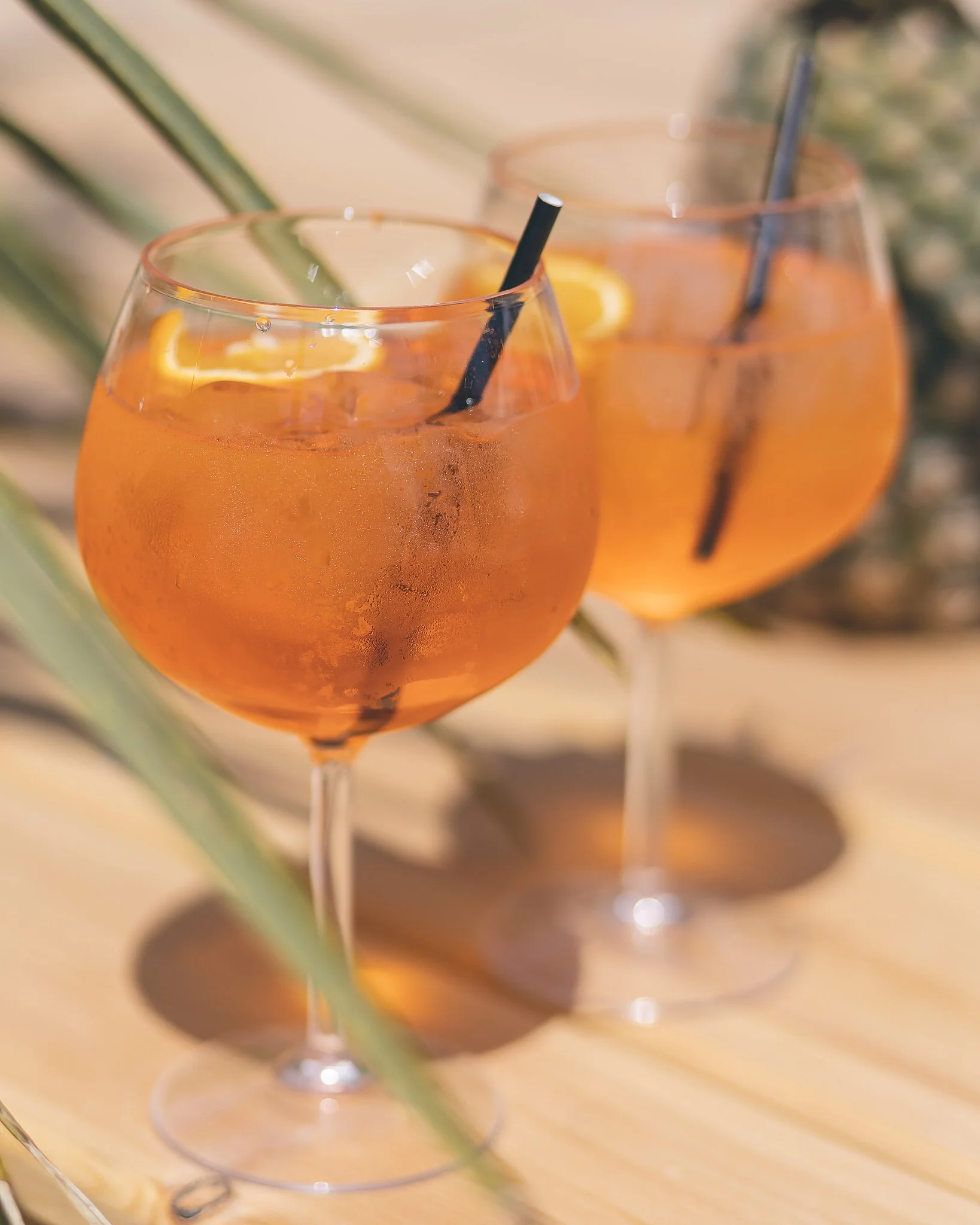 Two large wine glasses filled with orange-colored cocktail garnished with lemon slices, set on a wooden surface with green leaves in the background.
