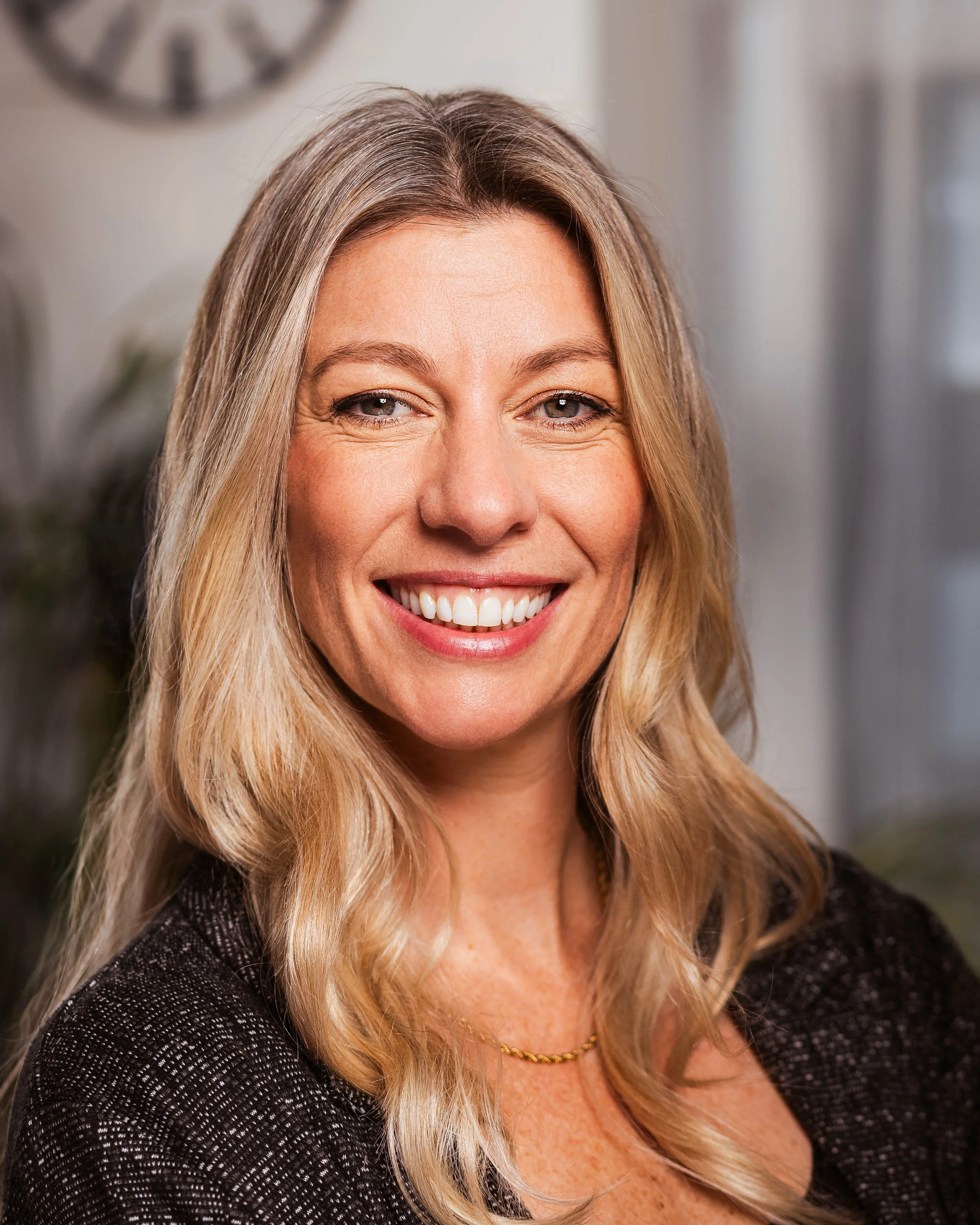 A woman with blonde, wavy hair and a broad smile, wearing a dark, sparkly top and a gold necklace in an indoor setting.