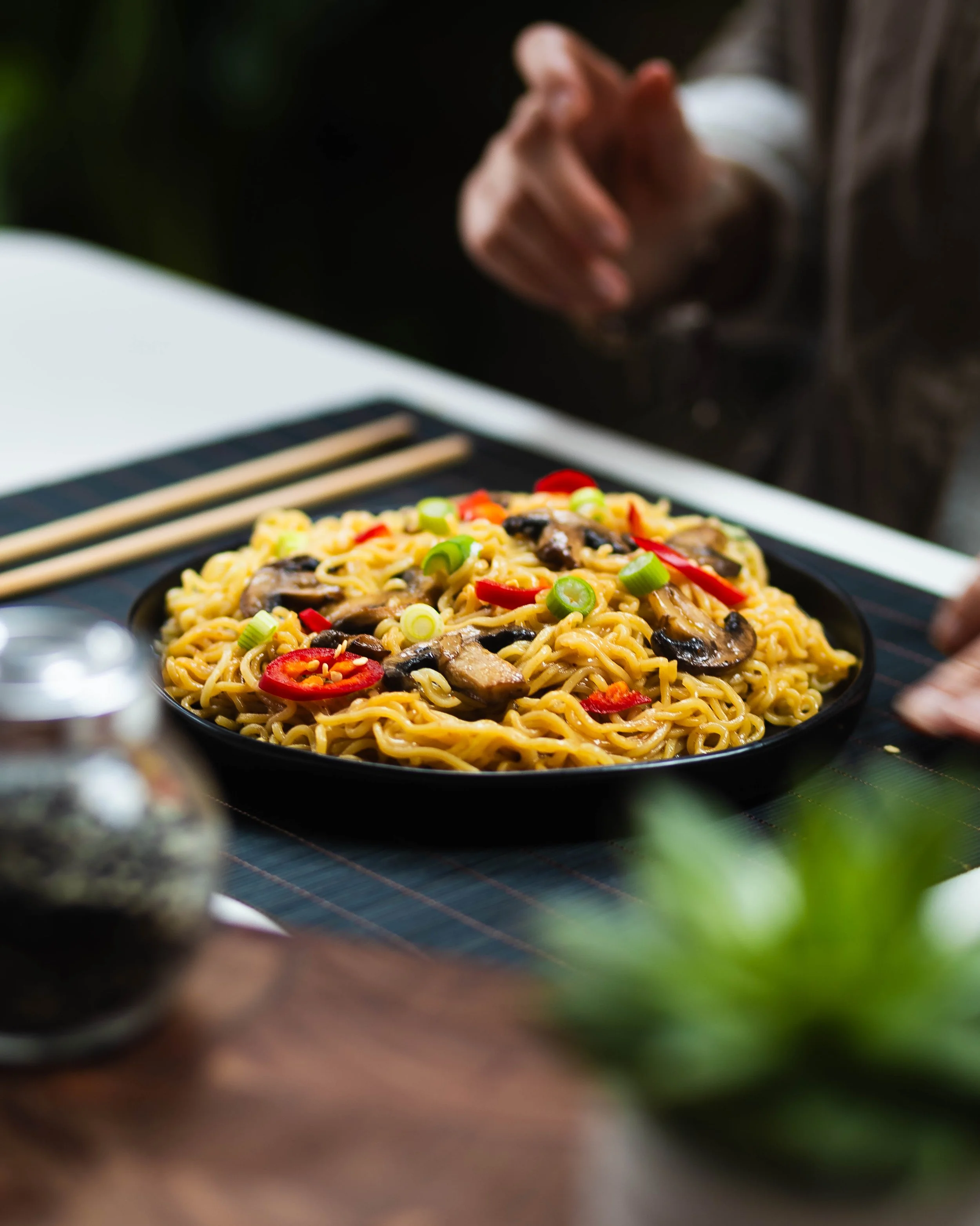 A plate of stir-fried noodles with mushrooms, sliced red chili peppers, and green onions on a table with chopsticks.