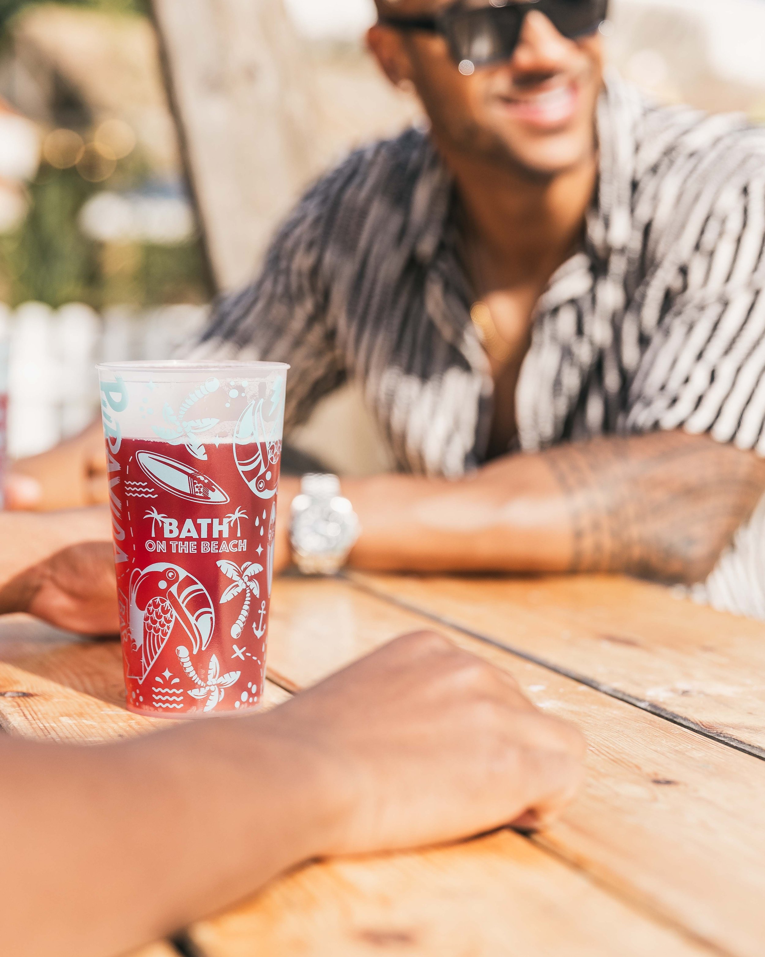 Lifestyle product photo of a cocktail in a 'Bath on the Beach' glass being enjoyed by two men at a sunny beach bar