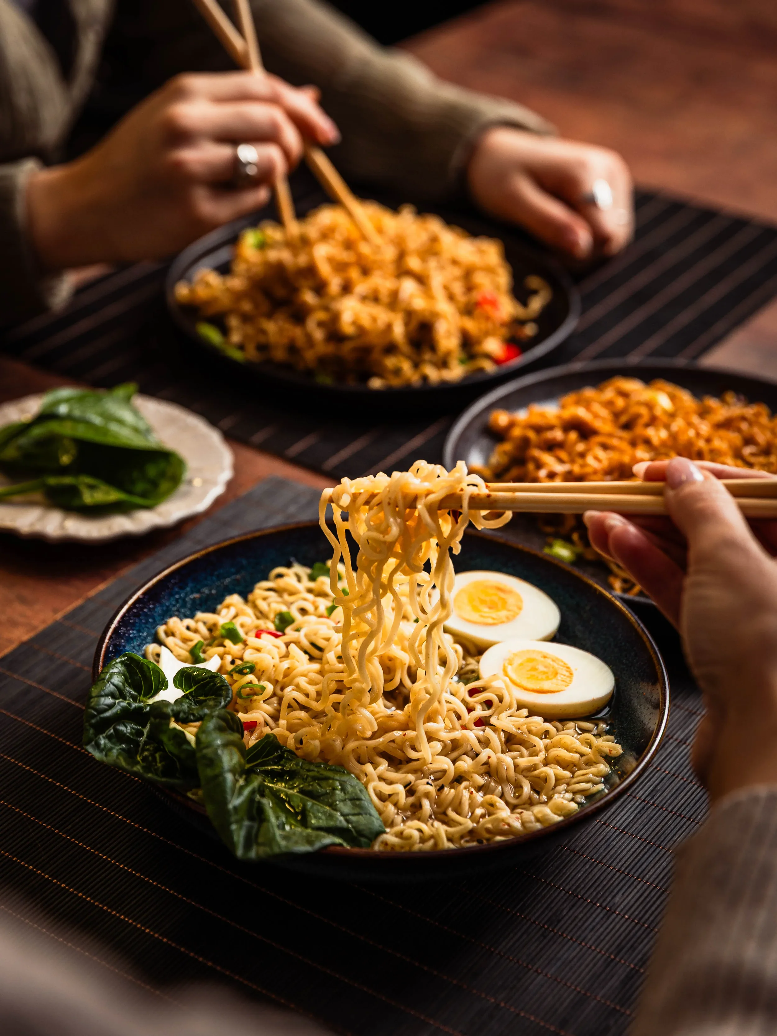 People enjoying bowls of ramen noodles with boiled eggs, leafy greens, and various Asian dishes at a dining table.