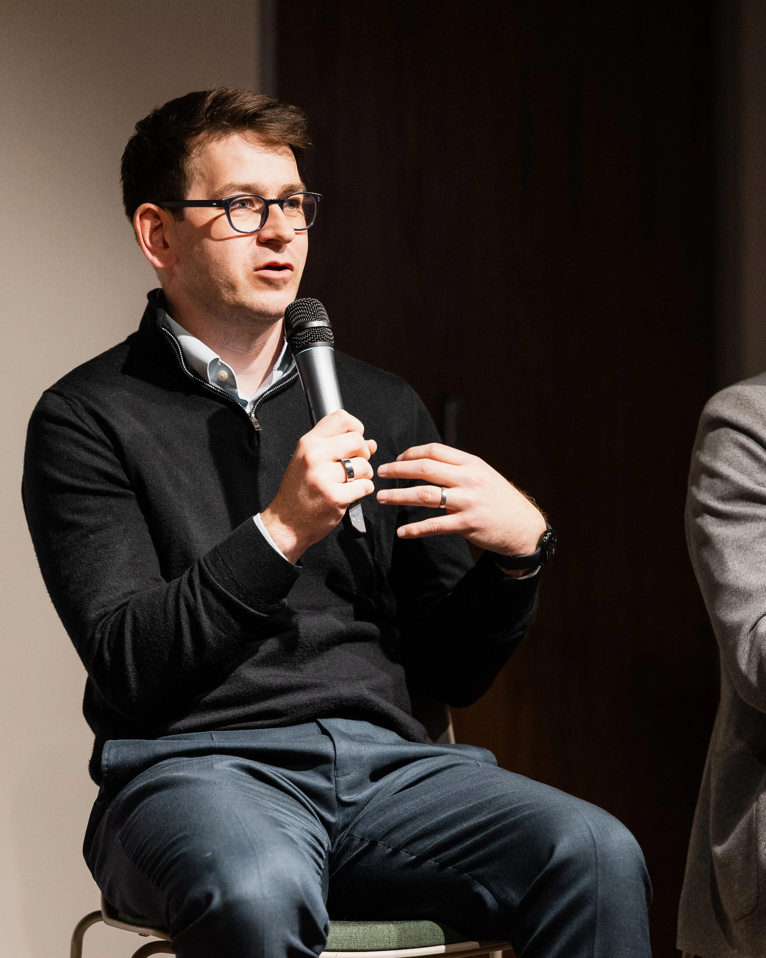 A smartly dressed man with glasses talking into a microphone on stage at a business presentation by London Photographer Bobby Shoebotham