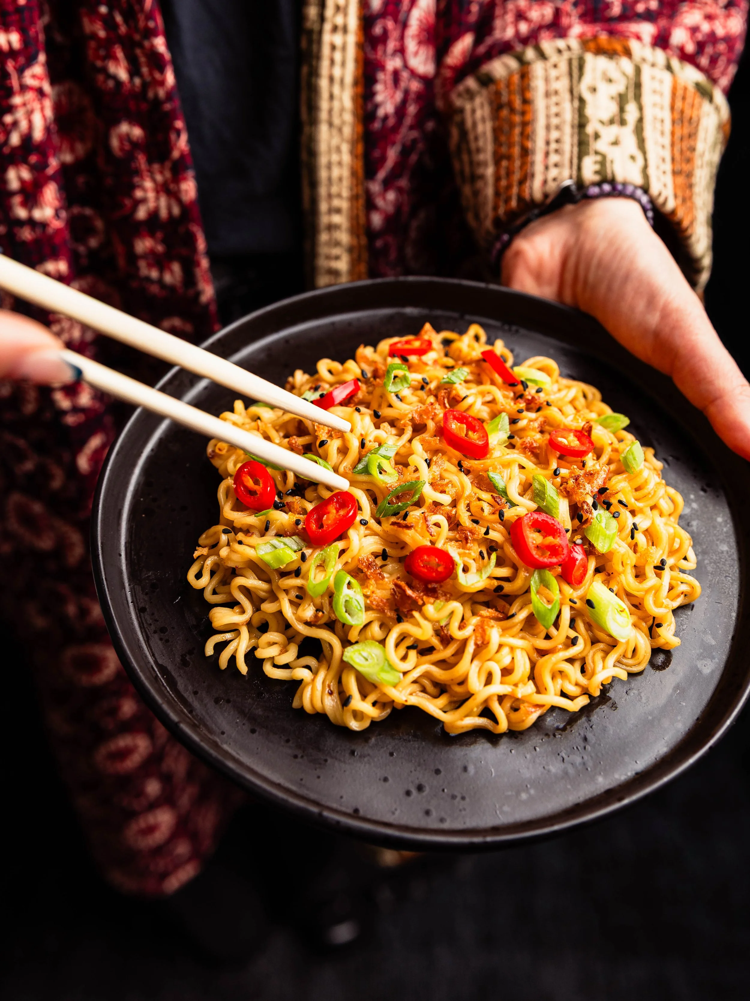 Close-up of a black bowl filled with cooked ramen noodles garnished with sliced red chili peppers, chopped green onions, and black sesame seeds, held by a person wearing a patterned jacket.