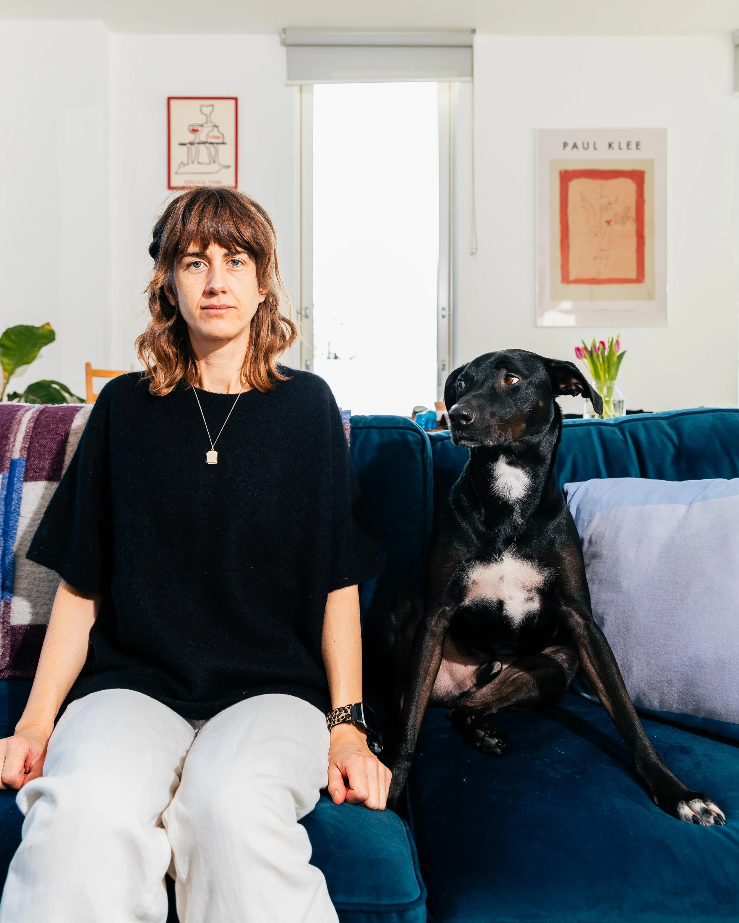 A woman with shoulder-length brown hair and a black sweater sitting on a blue sofa next to a large black dog with a white patch on its chest in a living room. The room has a white wall, two framed posters, and a sliding door with a blind in the backg
