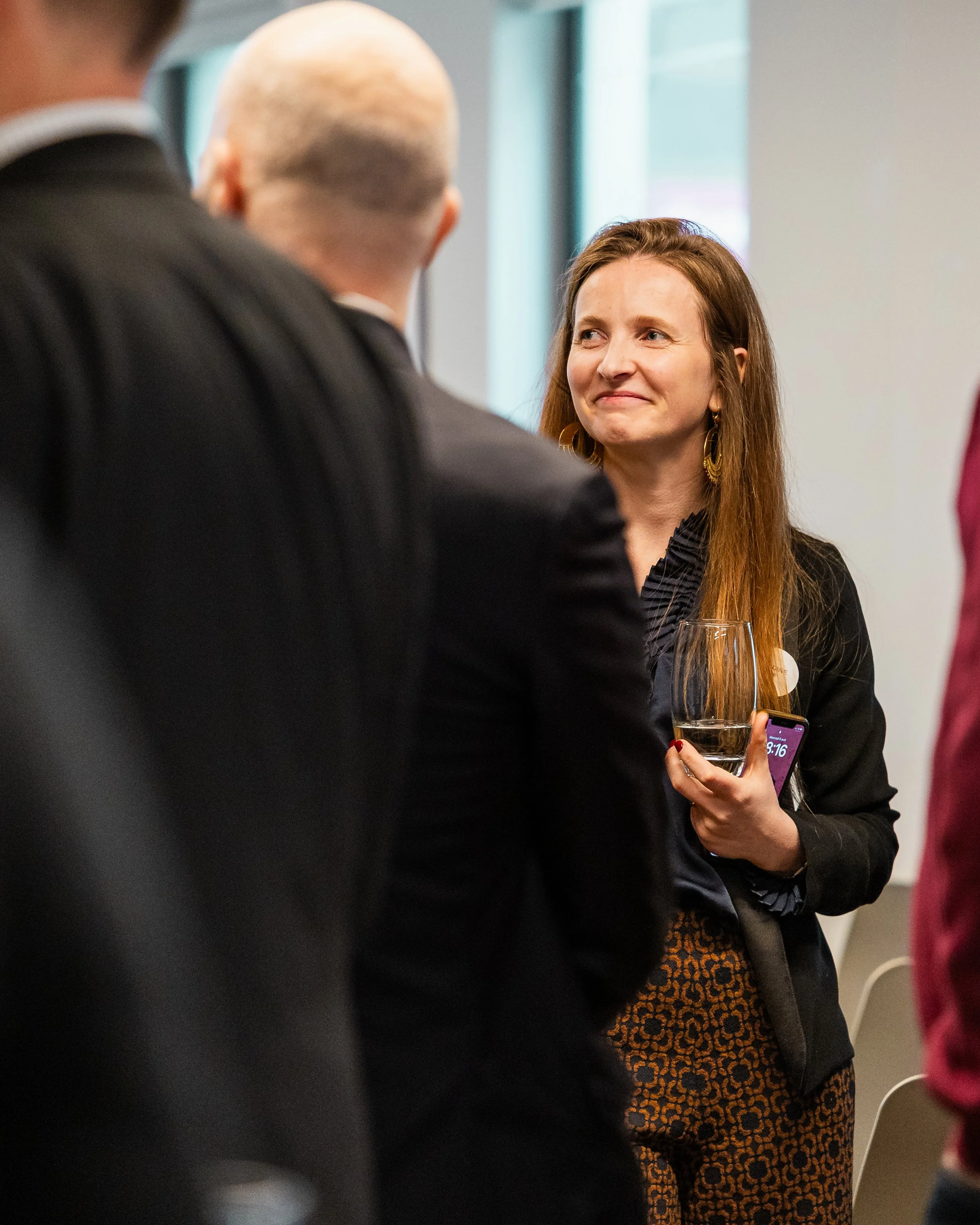 A woman holding a glass of wine and smiling at someone off camera at a business event by London Photographer Bobby Shoebotham