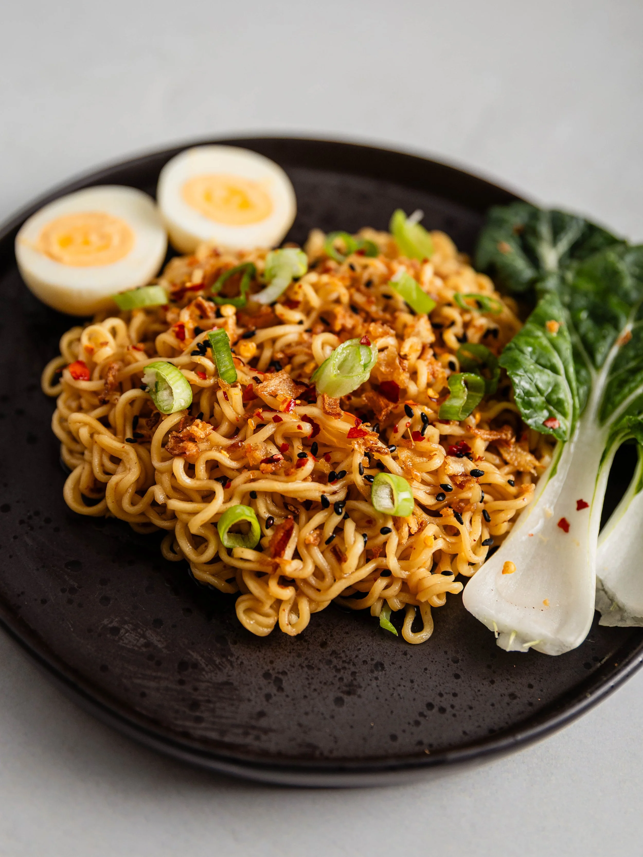 A plate of cooked ramen noodles garnished with chopped green onions, black sesame seeds, and red pepper flakes, with sliced boiled eggs and greens on the side.