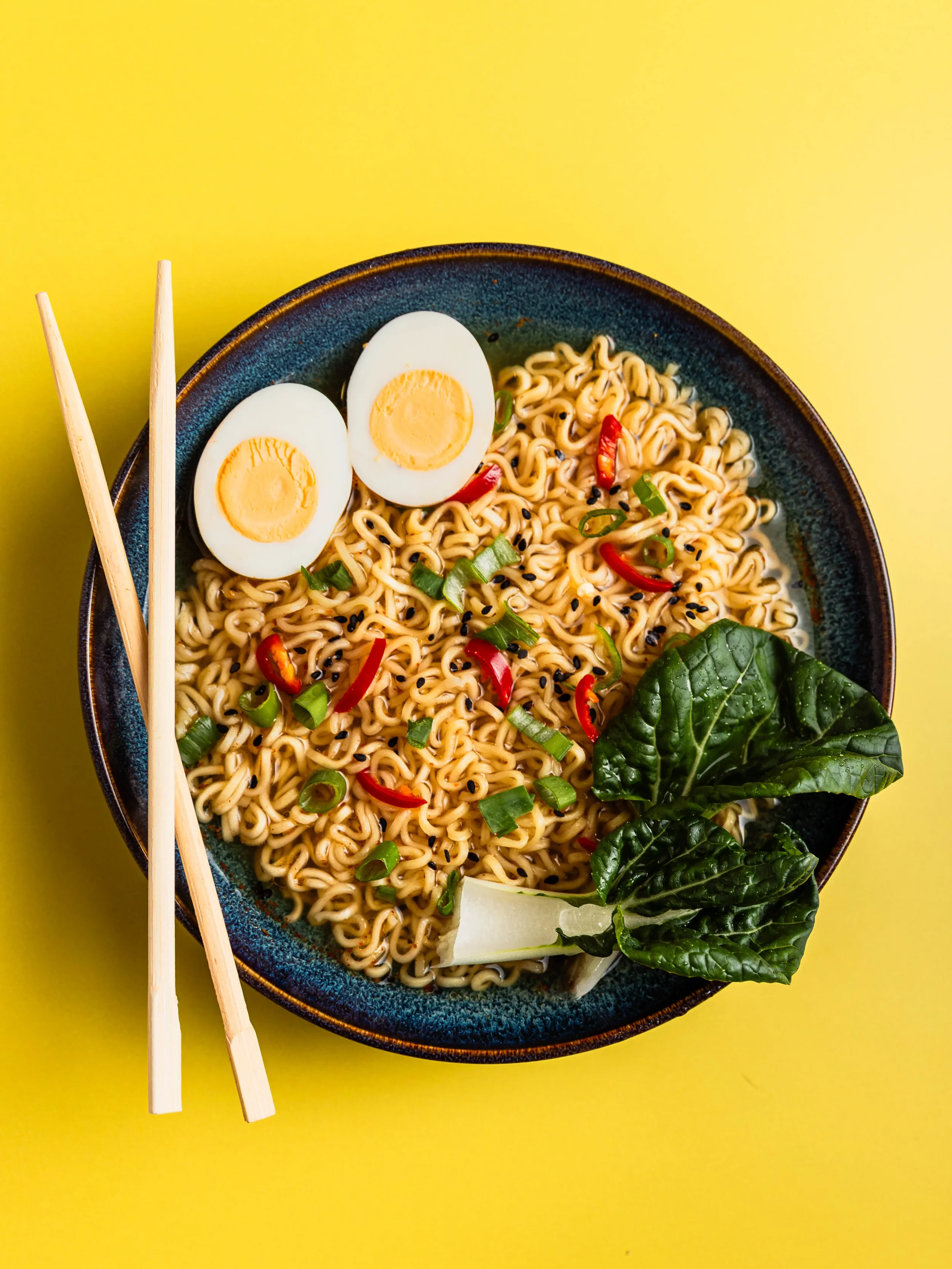 A bowl of ramen noodles topped with halved boiled eggs, chopped green onions, thin slices of red chili, black sesame seeds, and fresh spinach leaves on a yellow background, with chopsticks on the side.