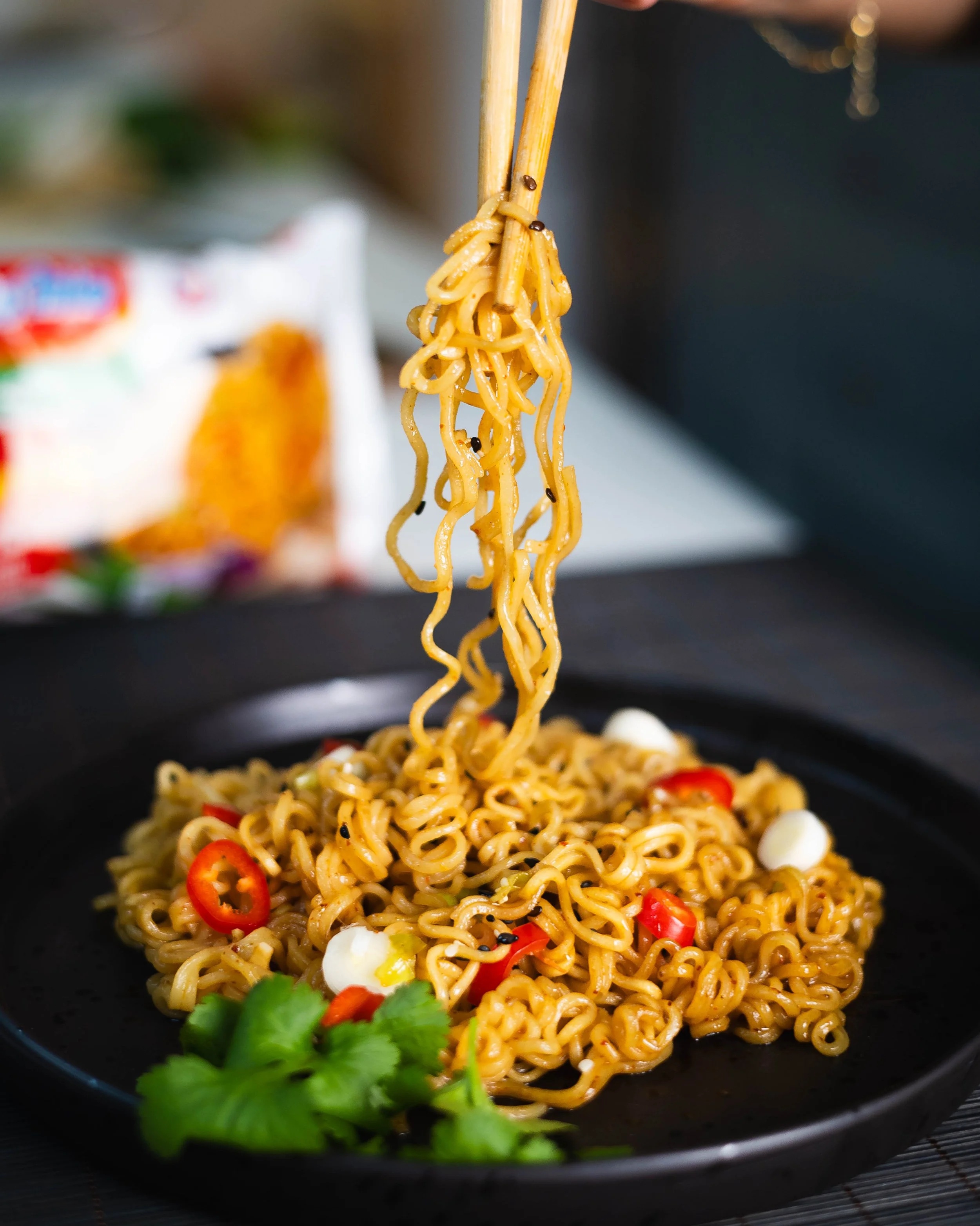 Professional product photograph of some noodles on a dark plate with some being held up by some chopsticks