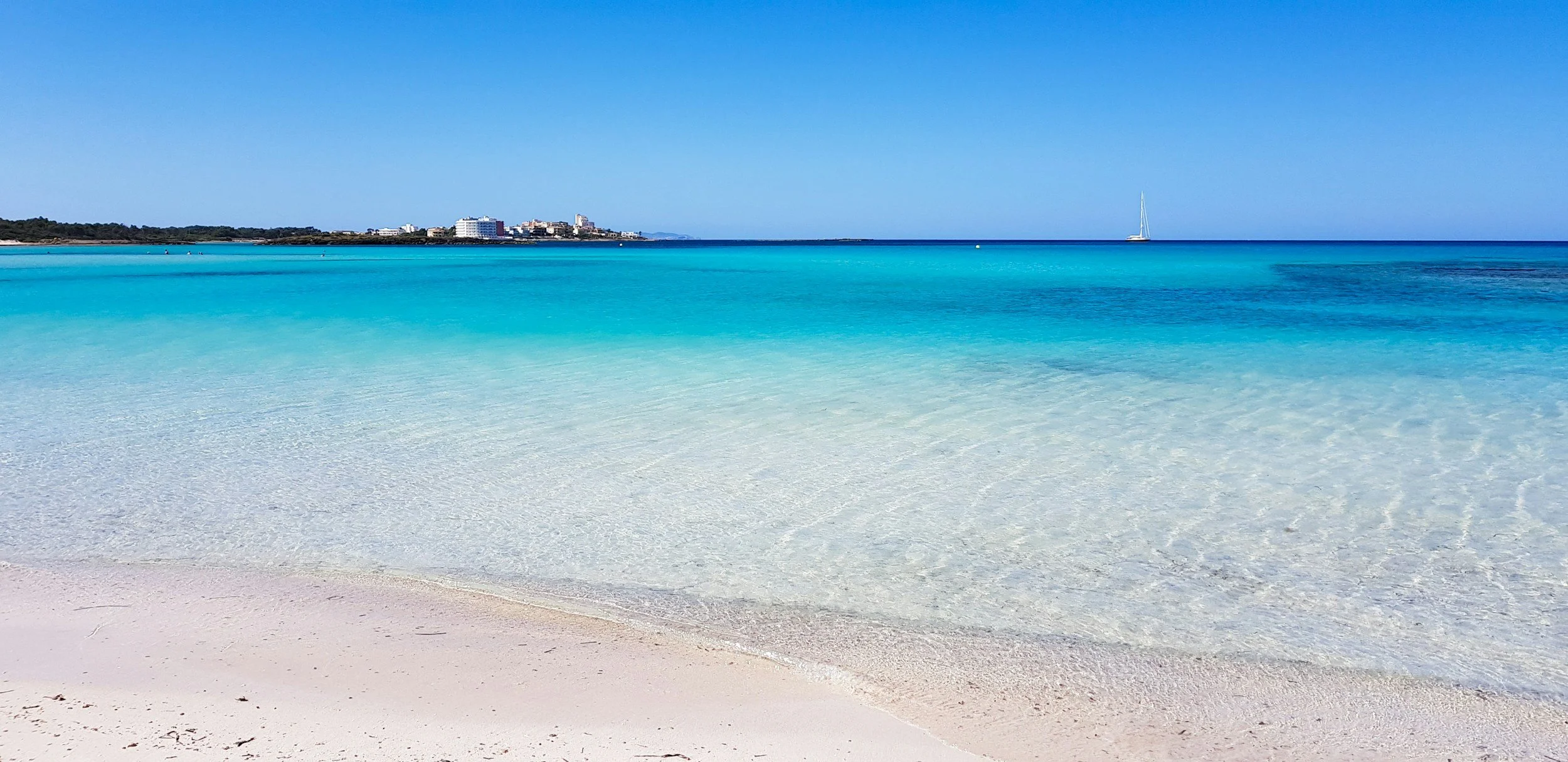 Clear turquoise ocean water meeting a white sandy beach with a distant city skyline and a sailboat on the horizon.