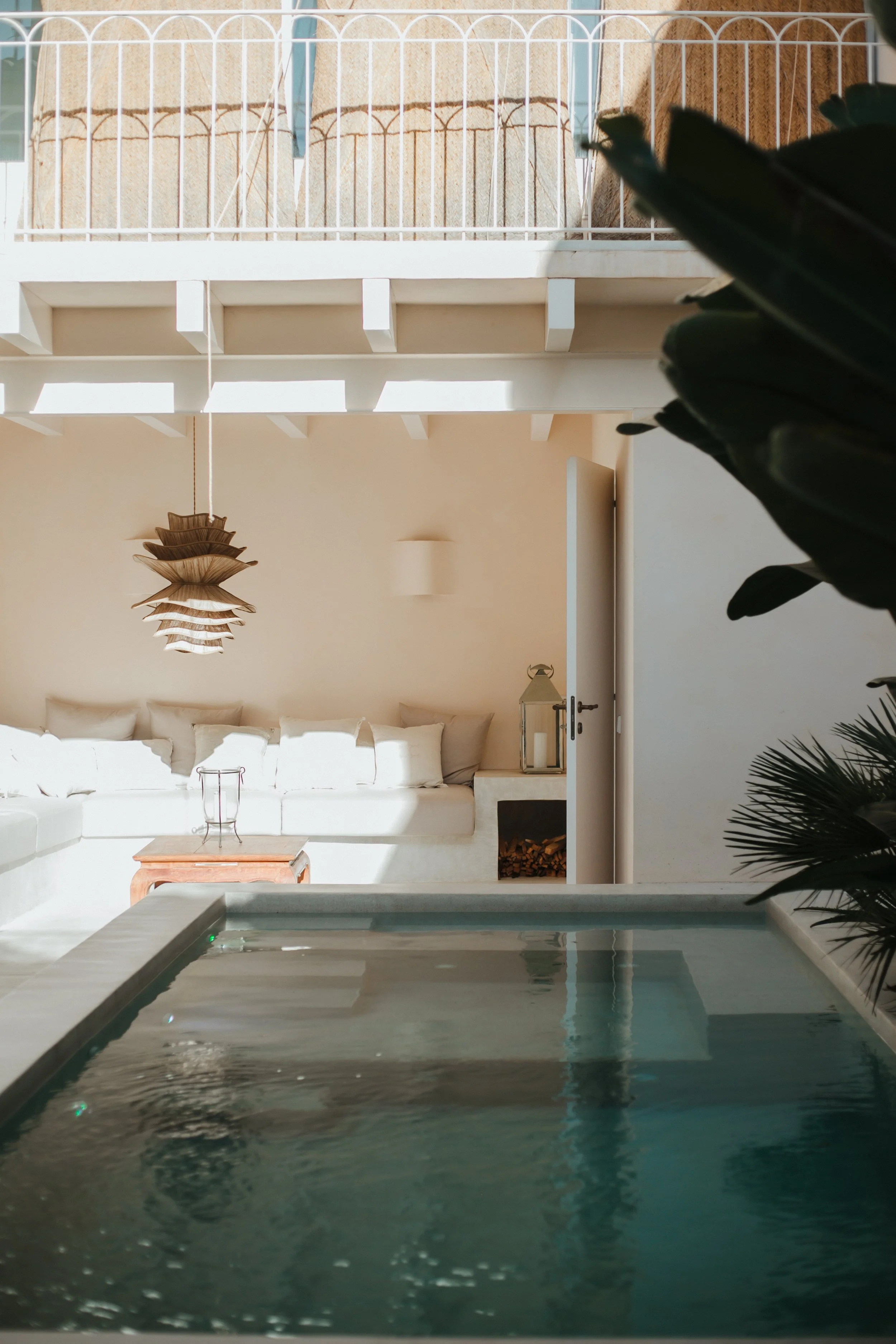 Indoor pool area with lounge chairs, a sofa with pillows, a small wooden table, and plant foliage in the foreground. Natural light enters the space, creating a relaxed atmosphere.