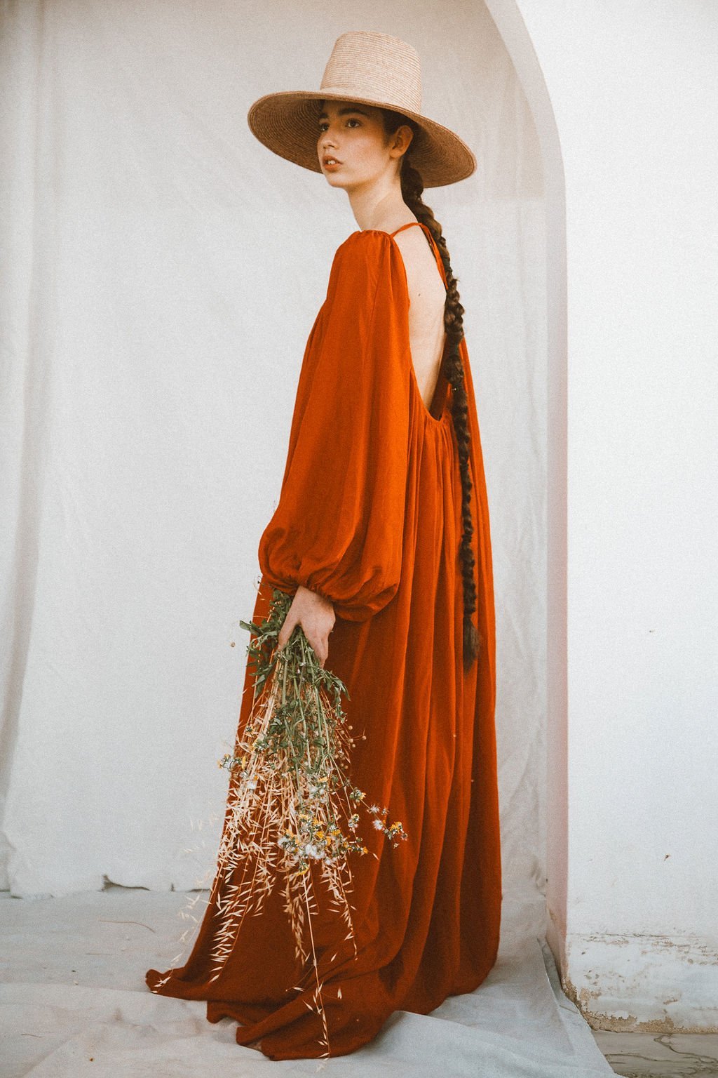 Woman wearing a wide-brimmed hat and a flowing red dress with an open back, holding a bouquet of wildflowers, standing against a white wall.