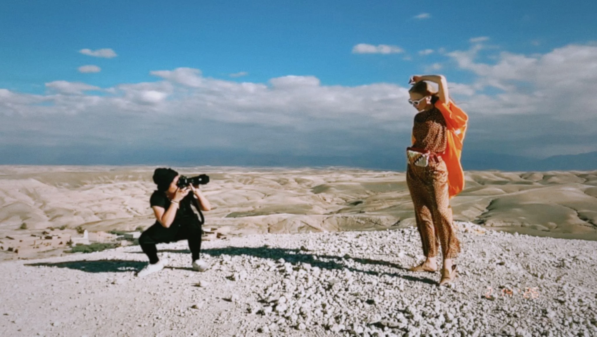 A woman dressed in a leopard-print outfit and orange cover-up is posing on a rocky, white terrain at a beach or desert. A photographer crouches down, taking her picture with a camera. The sky is blue with scattered clouds and the landscape stretches into the distance.