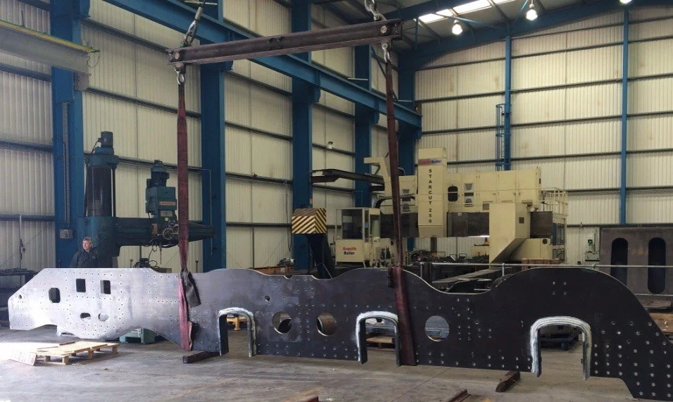 Large steam locomotive section in a factory, suspended from a crane, with industrial machinery and a worker in the background.