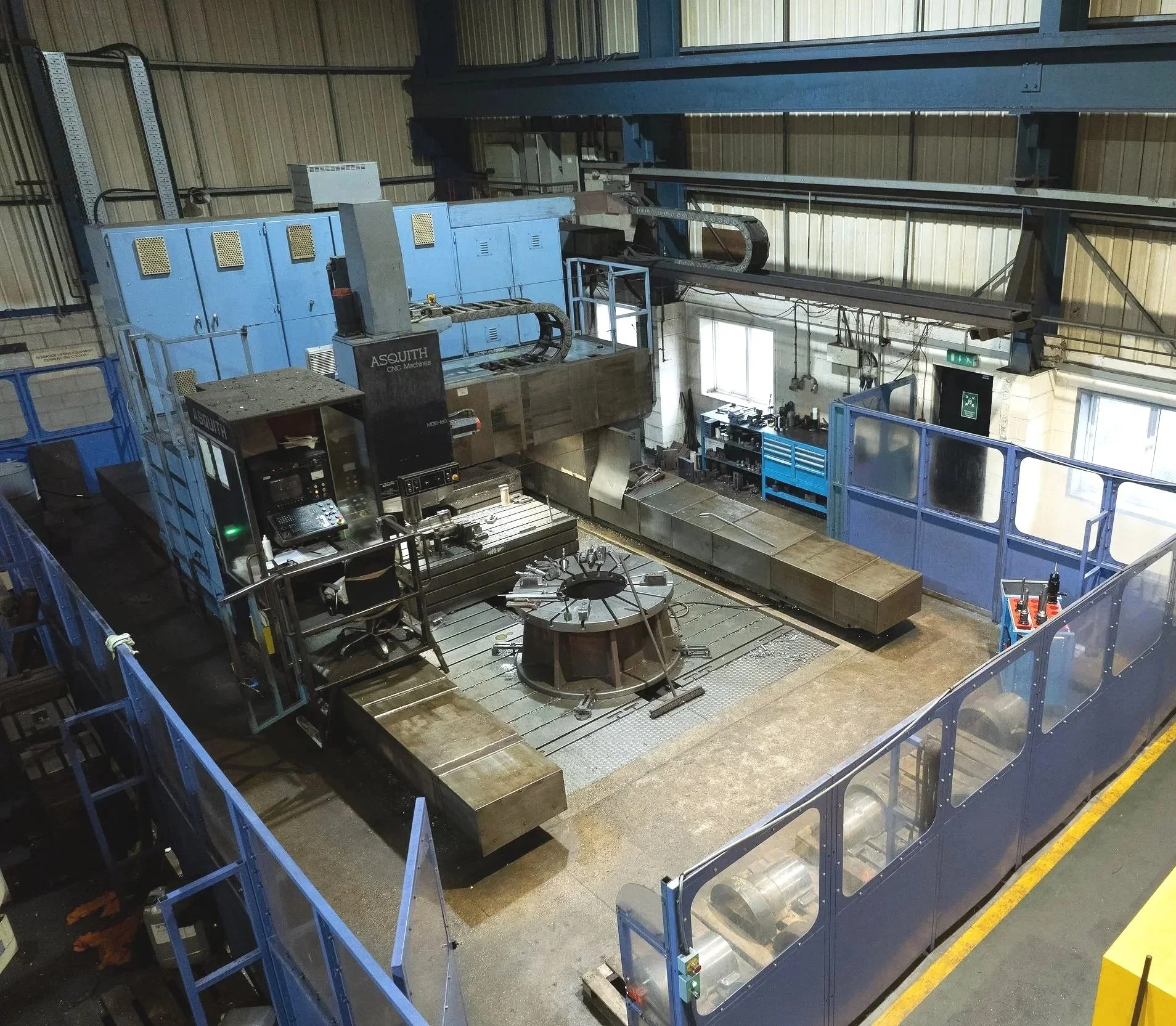 Industrial workshop featuring a large CNC machine with a central workpiece, surrounded by safety barriers and tools, within a spacious factory setting.