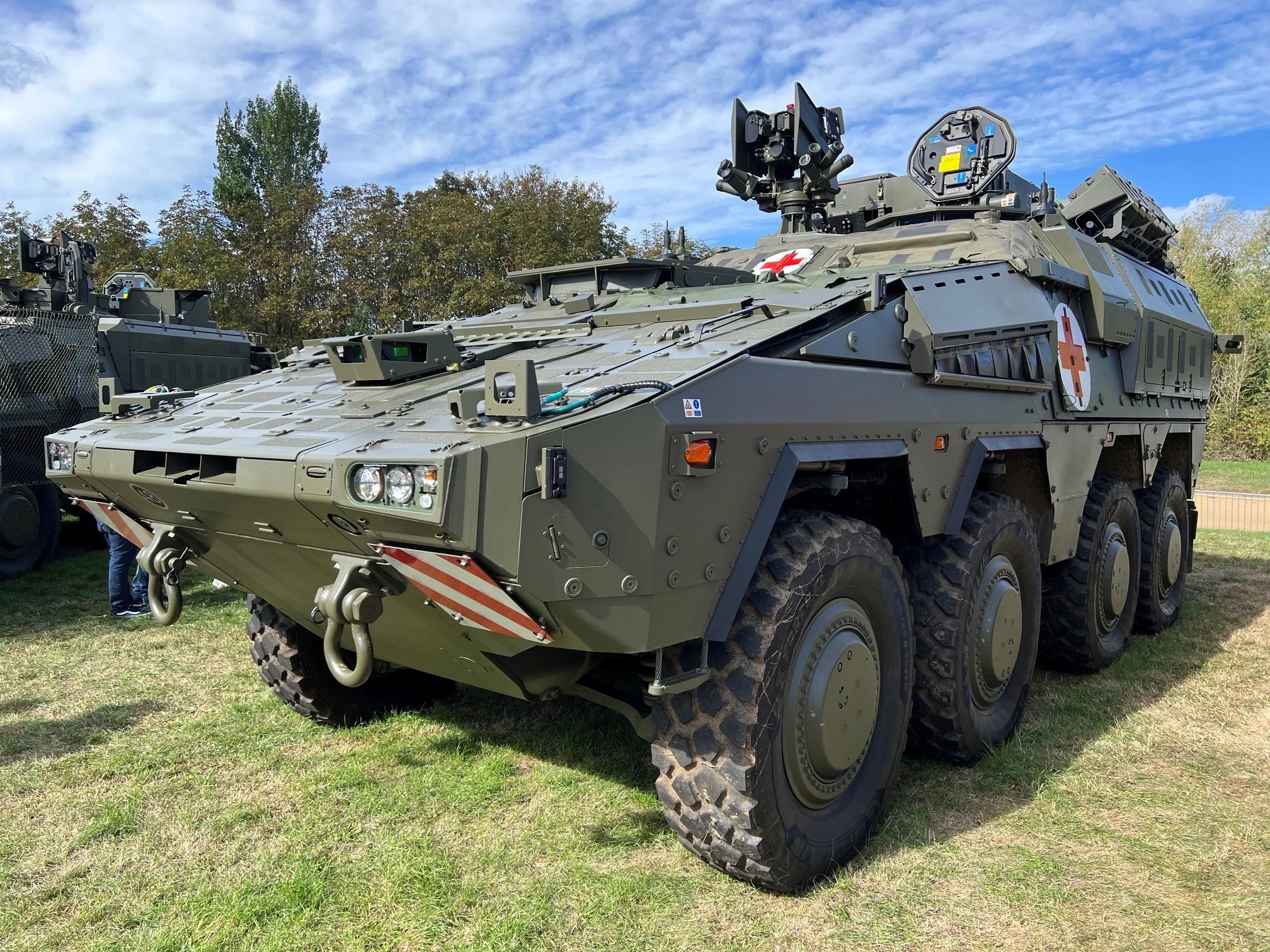 A military tank driving on a dirt road through a wooded area with green trees and grass.