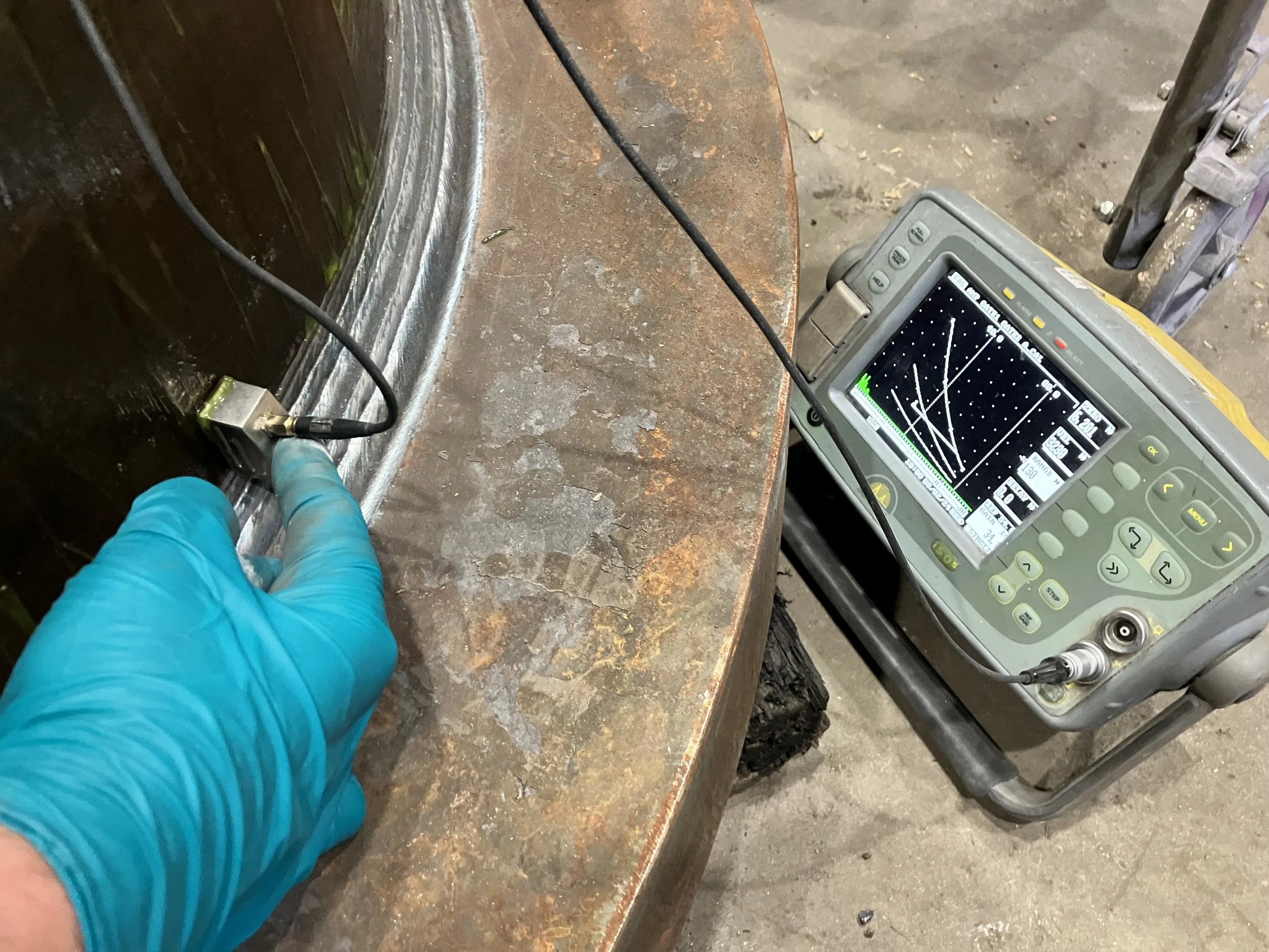 A worker wearing a blue glove is testing or measuring a metal surface with a probe connected to a diagnostic equipment displaying a graph or wave pattern.