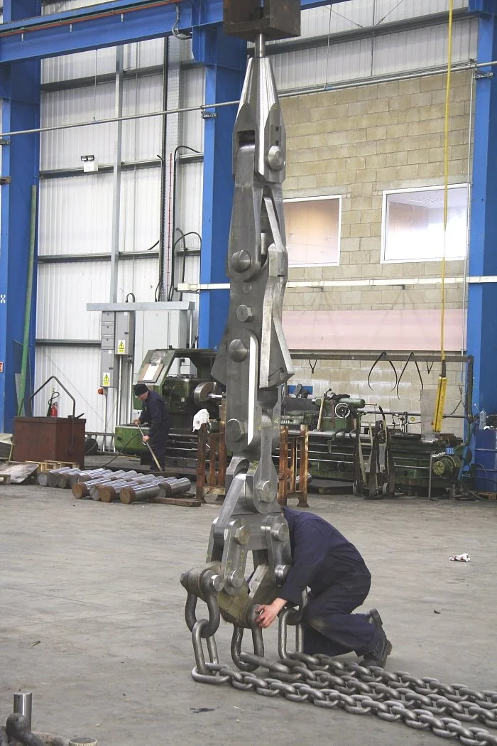A worker kneeling inside a factory or industrial setting, holding a large metal chain attached to a massive metal lifting component hanging from an overhead crane, with debris and equipment in the background.