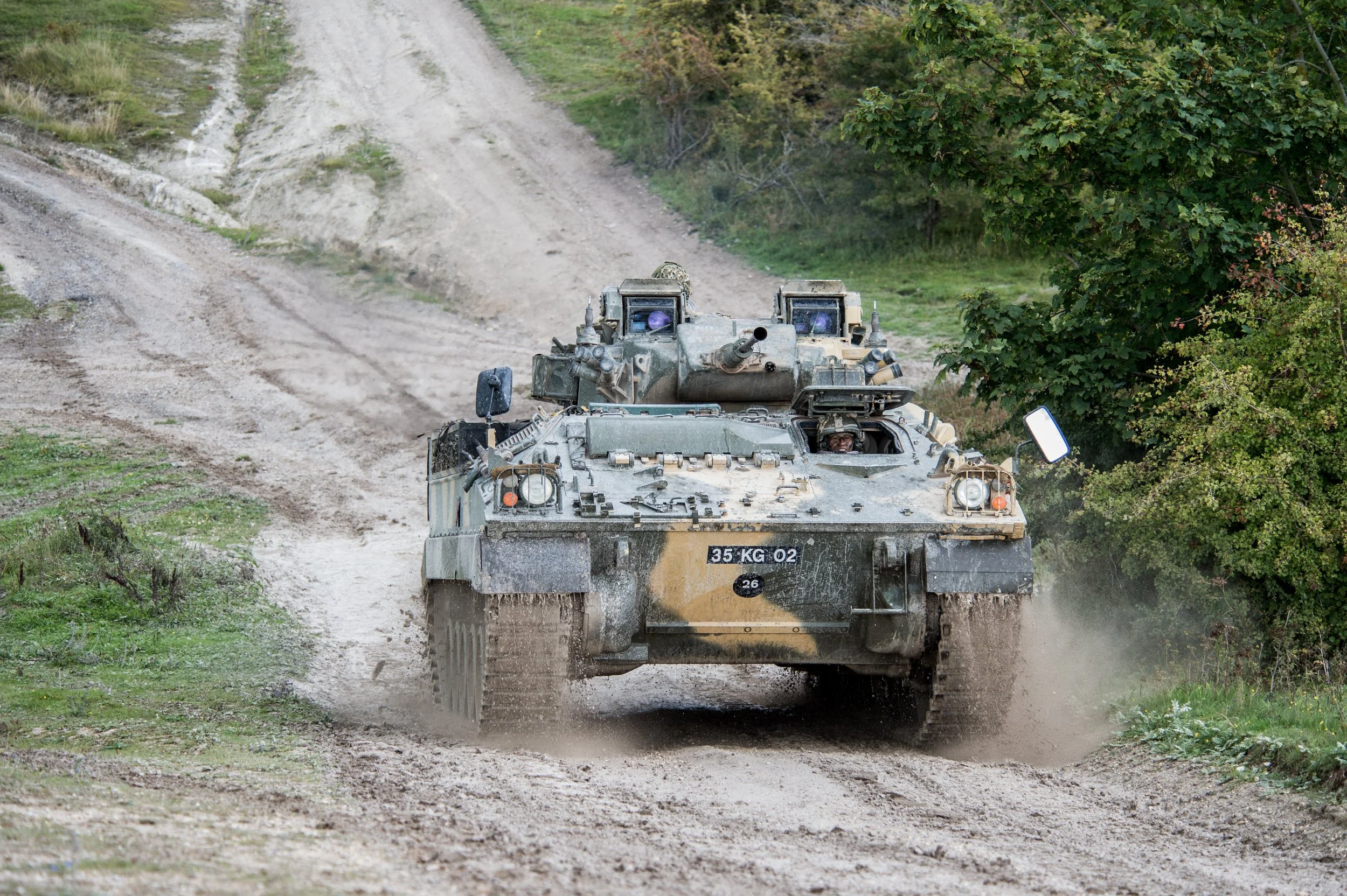 A military tank driving on a dirt road through a rural area with trees on the side.