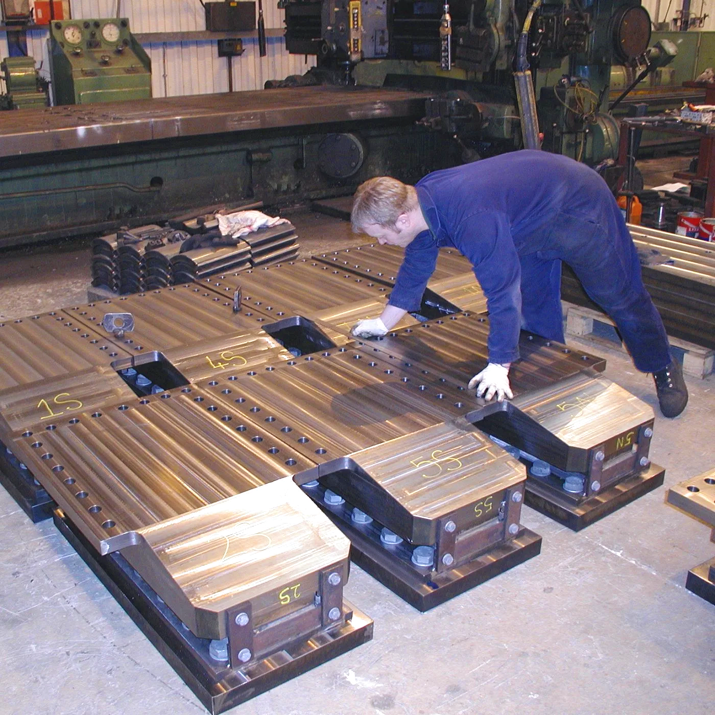 A worker in a blue coverall is assembling metal components on a factory floor to make large industrial parts, with machinery and tools visible in the background.