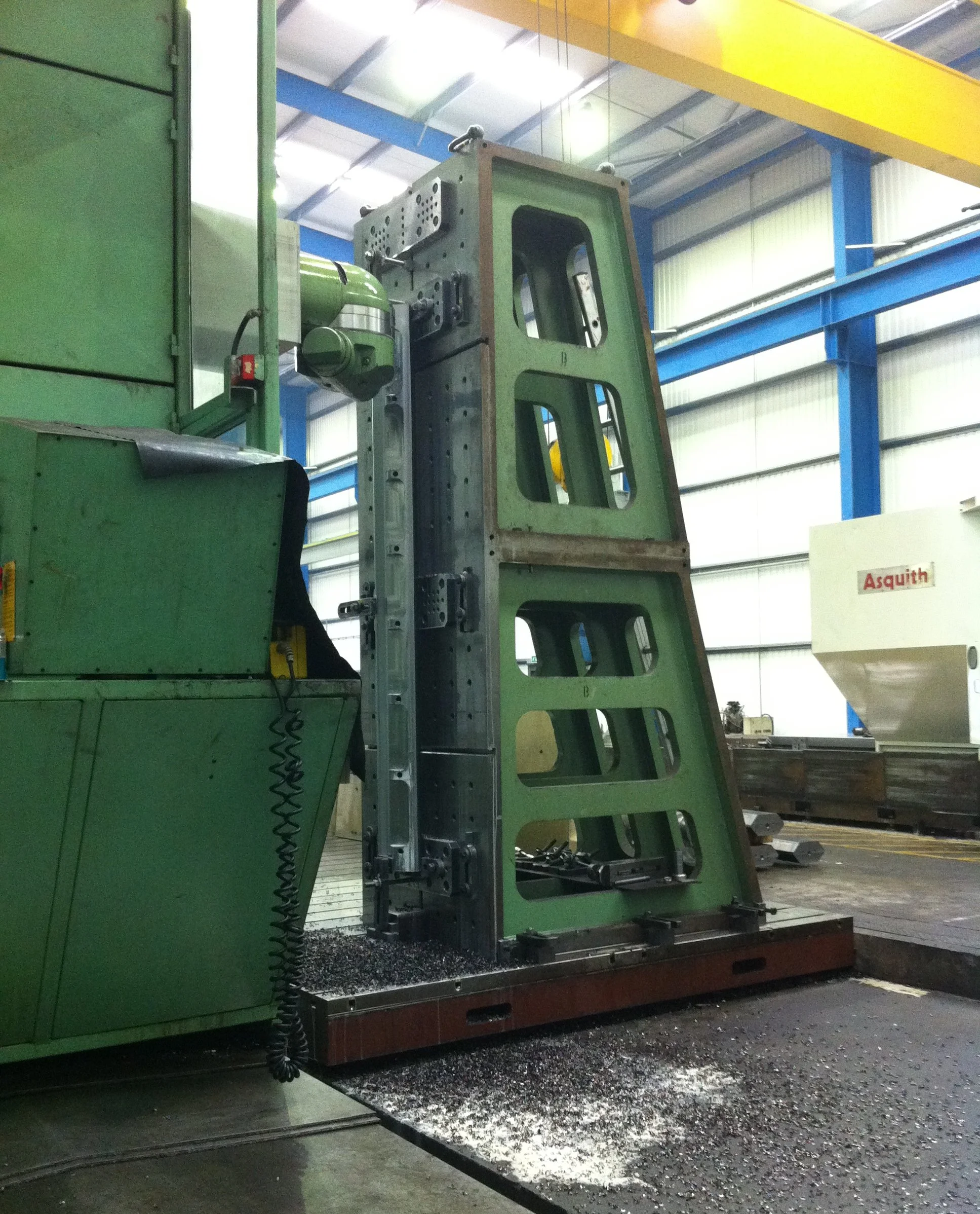 Large steel component being machined or assembled in an industrial factory, with a green machine and metal shavings on the floor.