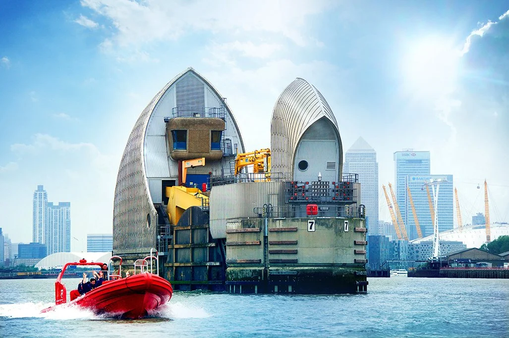 A series of modern, silver, shell-shaped buildings along a river with construction equipment, including yellow cranes, in front of them under a cloudy sky.