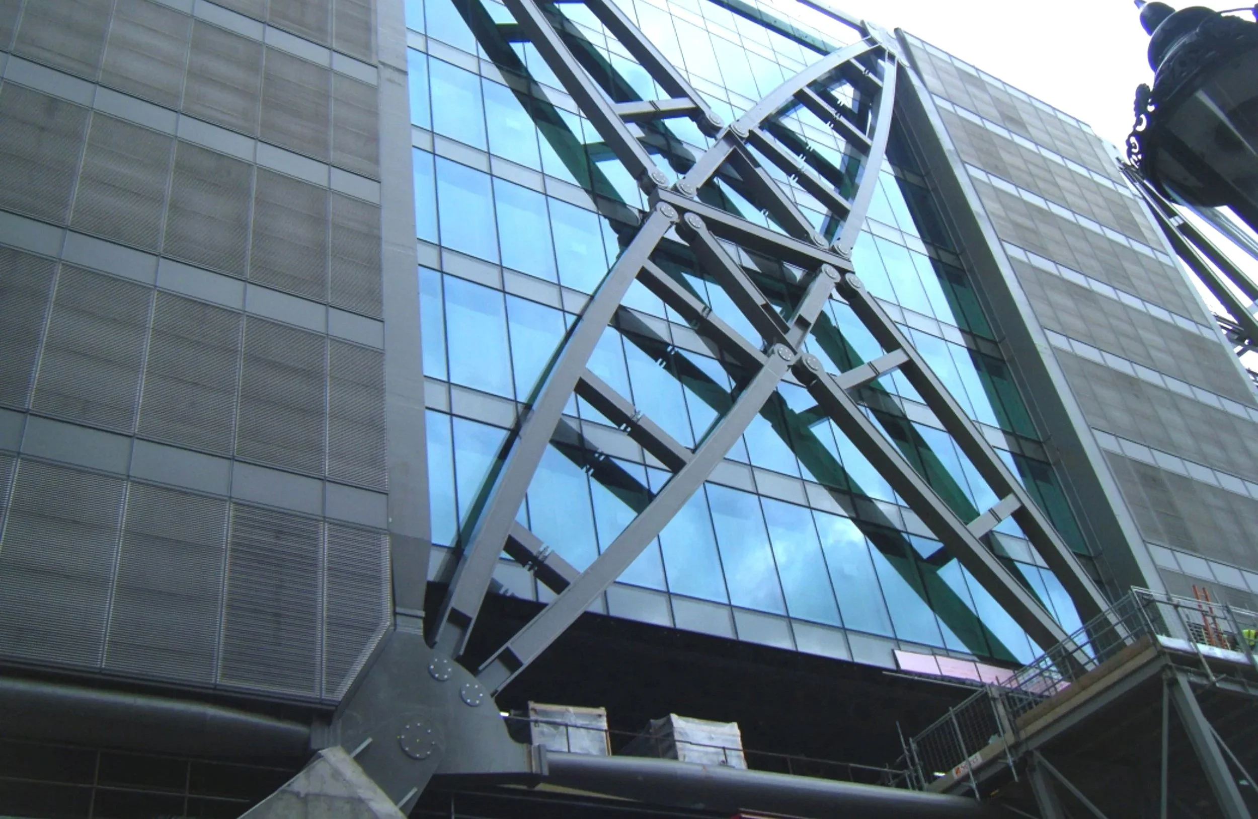 Close-up of a construction crane next to a modern glass building, with reflections of the sky and surrounding structures in the windows.
