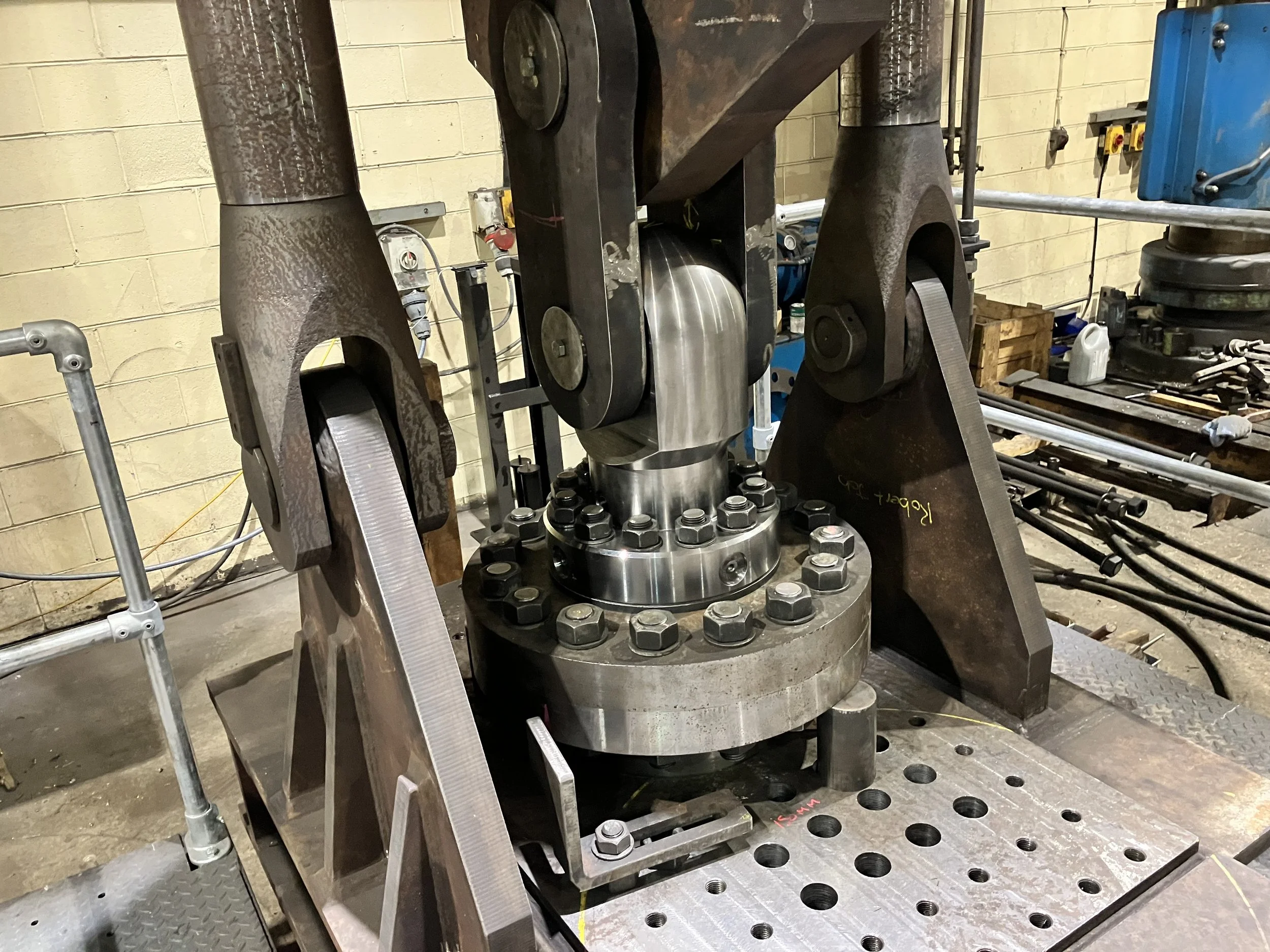 Close-up of a large industrial machine component with metal plates, bolts, and heavy-duty metal arms in a workshop setting.