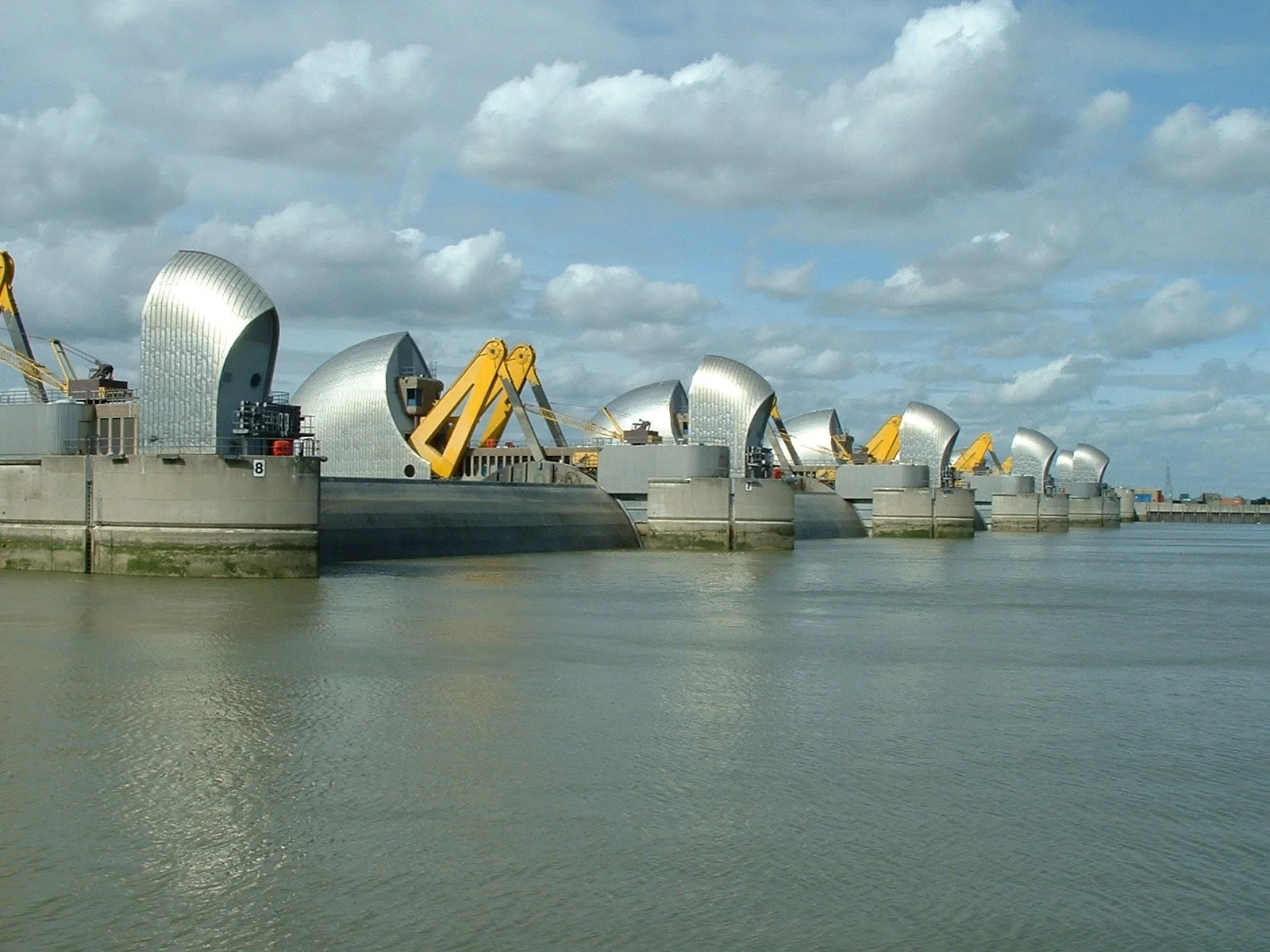 A series of modern, silver, shell-shaped buildings along a river with construction equipment, including yellow cranes, in front of them under a cloudy sky.
