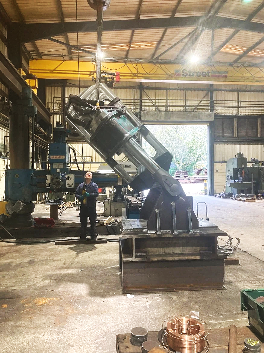 A worker in safety gear stands in an industrial workshop near a large robotic arm lifting a part. The workshop contains various machinery, metal parts, and tools, with an open garage door revealing outdoor sunlight.