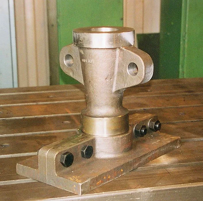 Metal mechanical part on a wooden surface, with bolt holes and a base, possibly a mounting bracket or connector.