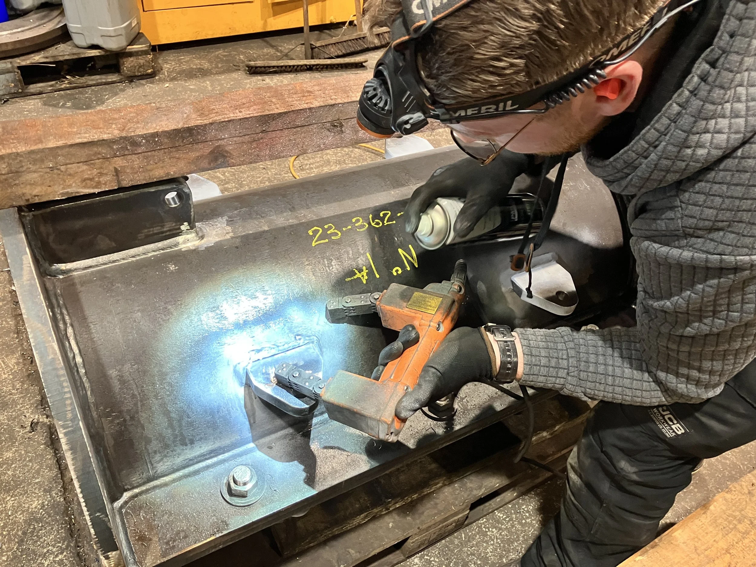 A worker welding a metal pipe with a handheld welding tool in a workshop, wearing safety glasses, gloves, and a headset, with yellow markings on the pipe.