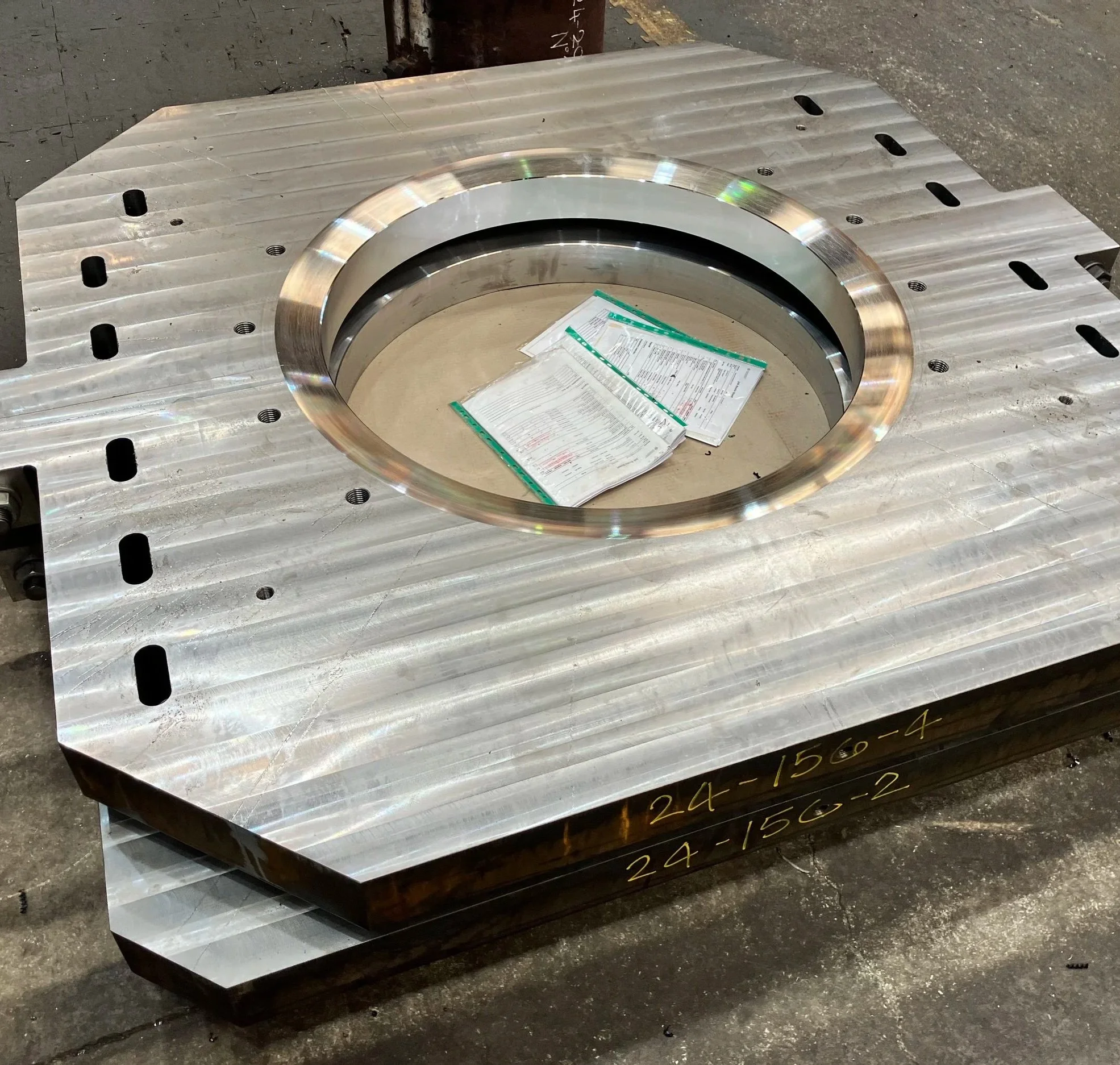Large metallic industrial part with a circular opening in the center, placed on a concrete floor with some paperwork on top.