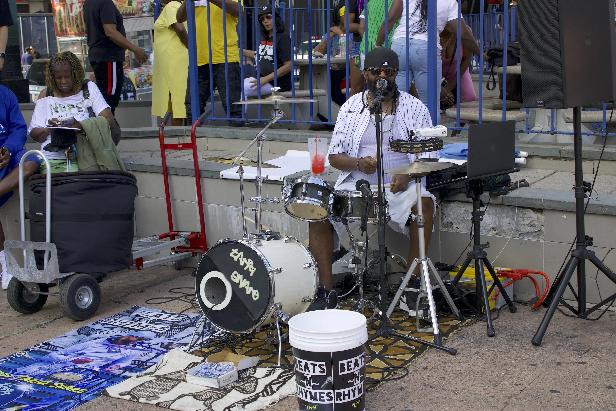 Street performer playing drums with a microphone, surrounded by people, equipment, and a "BEATS RHYMES" bucket on a sidewalk.