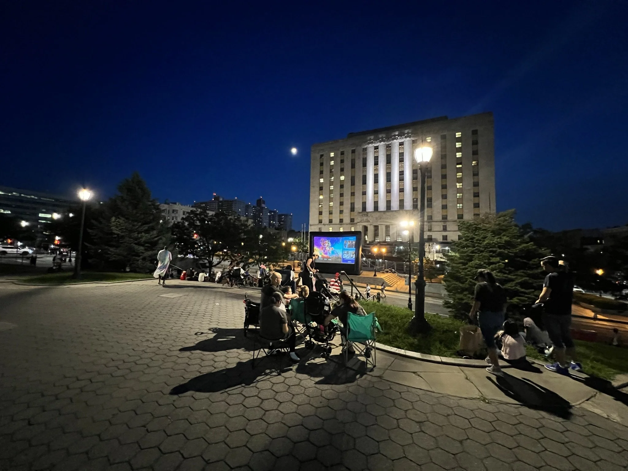 Outdoor movie screening at night in a park with people sitting in folding chairs, an illuminated building in the background, and a large screen displaying an animated film. The sky is dark, and streetlights are casting light.