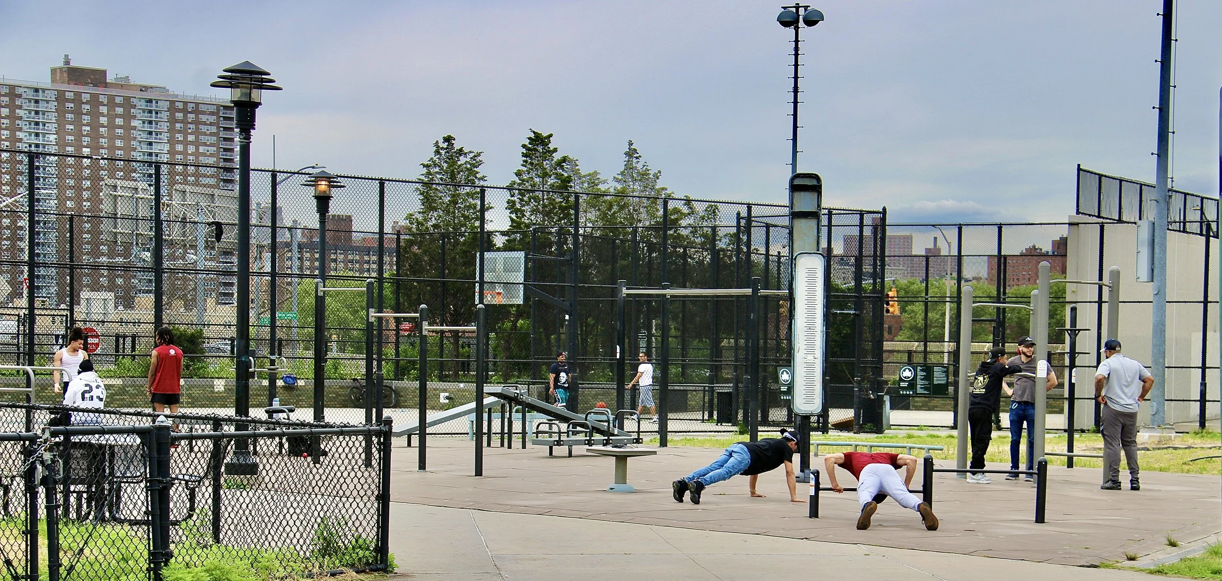 Outdoor fitness area with people exercising and socializing, urban background with buildings and trees, chain link fence and benches visible.