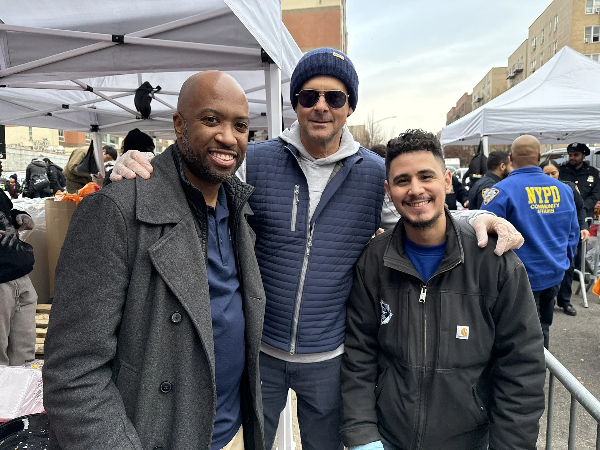 Three men smiling at a community event or charity gathering with tents and NYPD officers in the background.