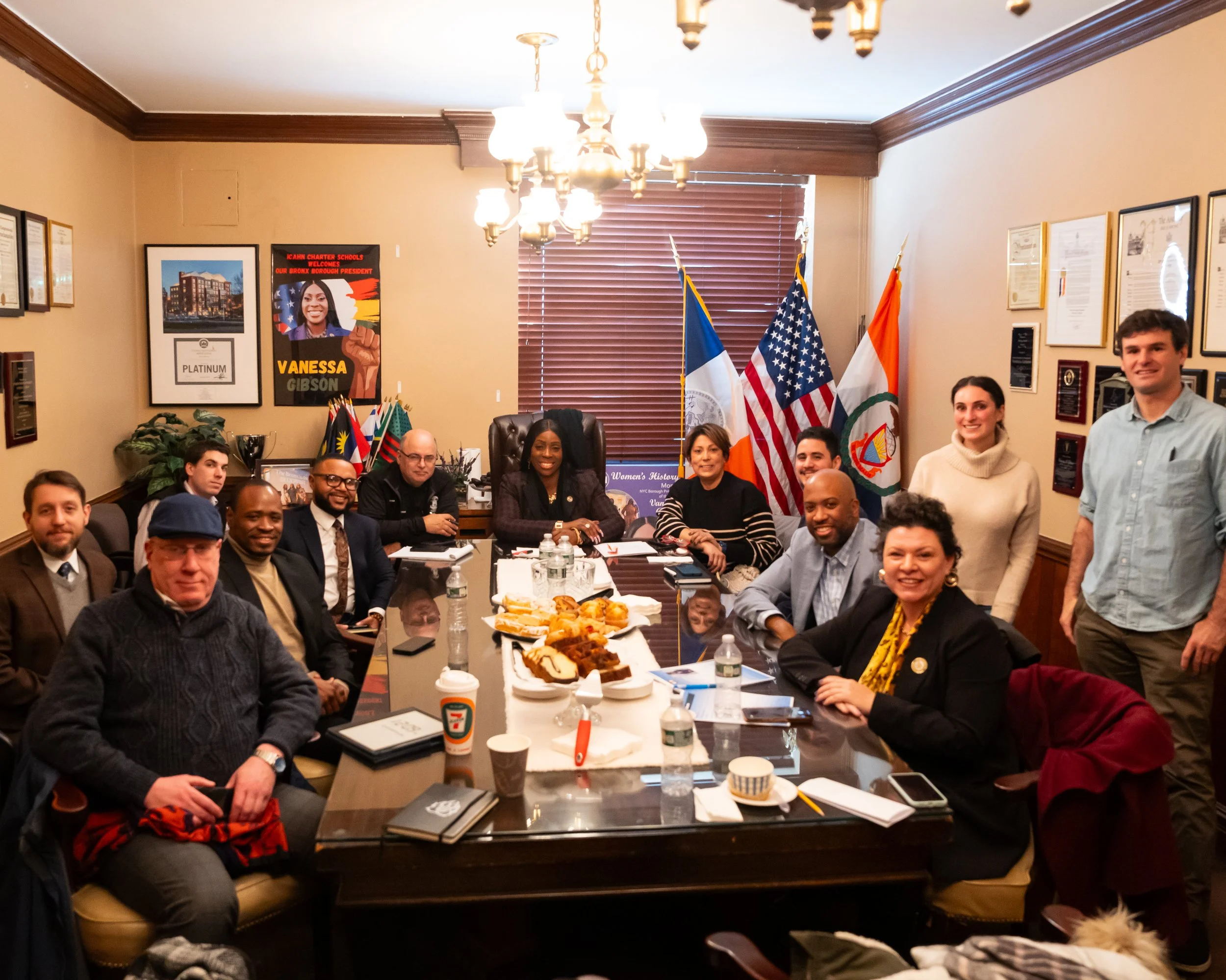 Group of people sitting around a conference table with flags and a poster on the wall, engaged in a formal meeting.