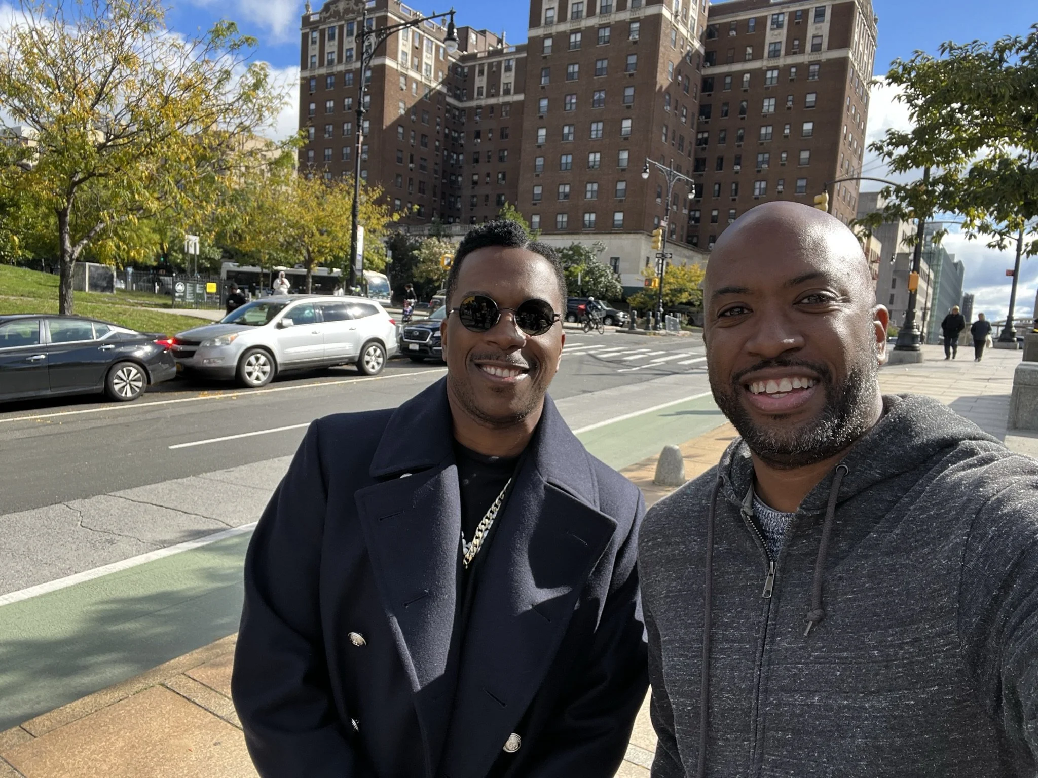 Two men smiling on a city street with a building and parked cars in the background.
