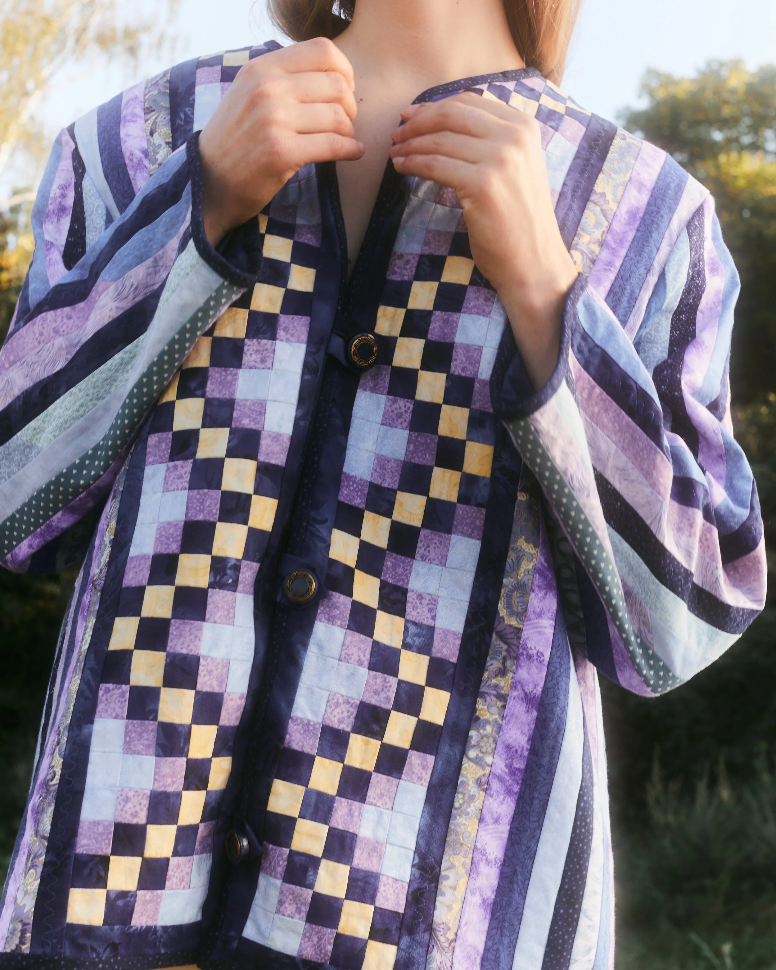 Close-up of a person adjusting a multicolored patchwork quilted garment outdoors with trees and sky in the background.