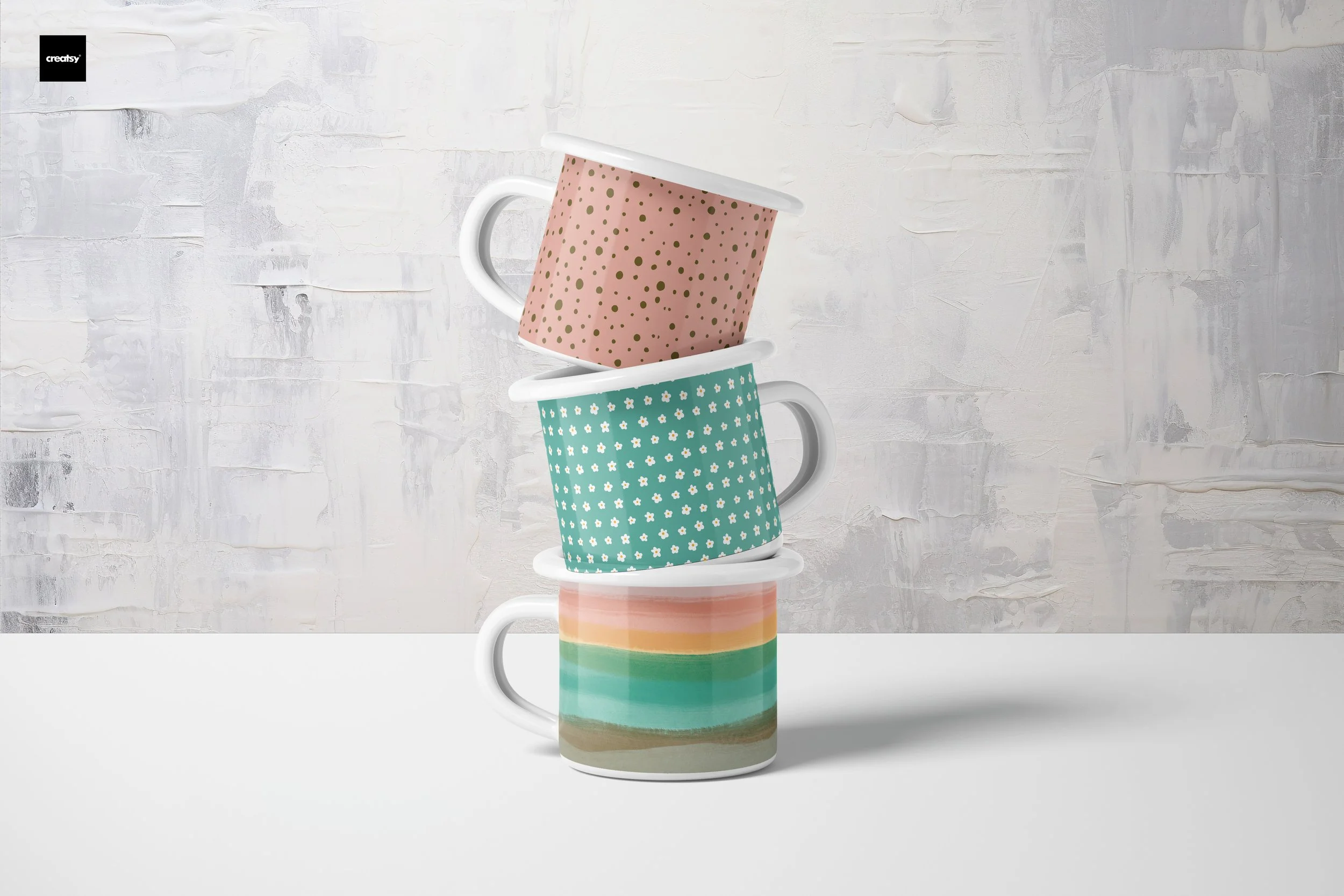 Three colorful ceramic mugs stacked on top of each other, with the top mug tilted, against a white and gray textured background.