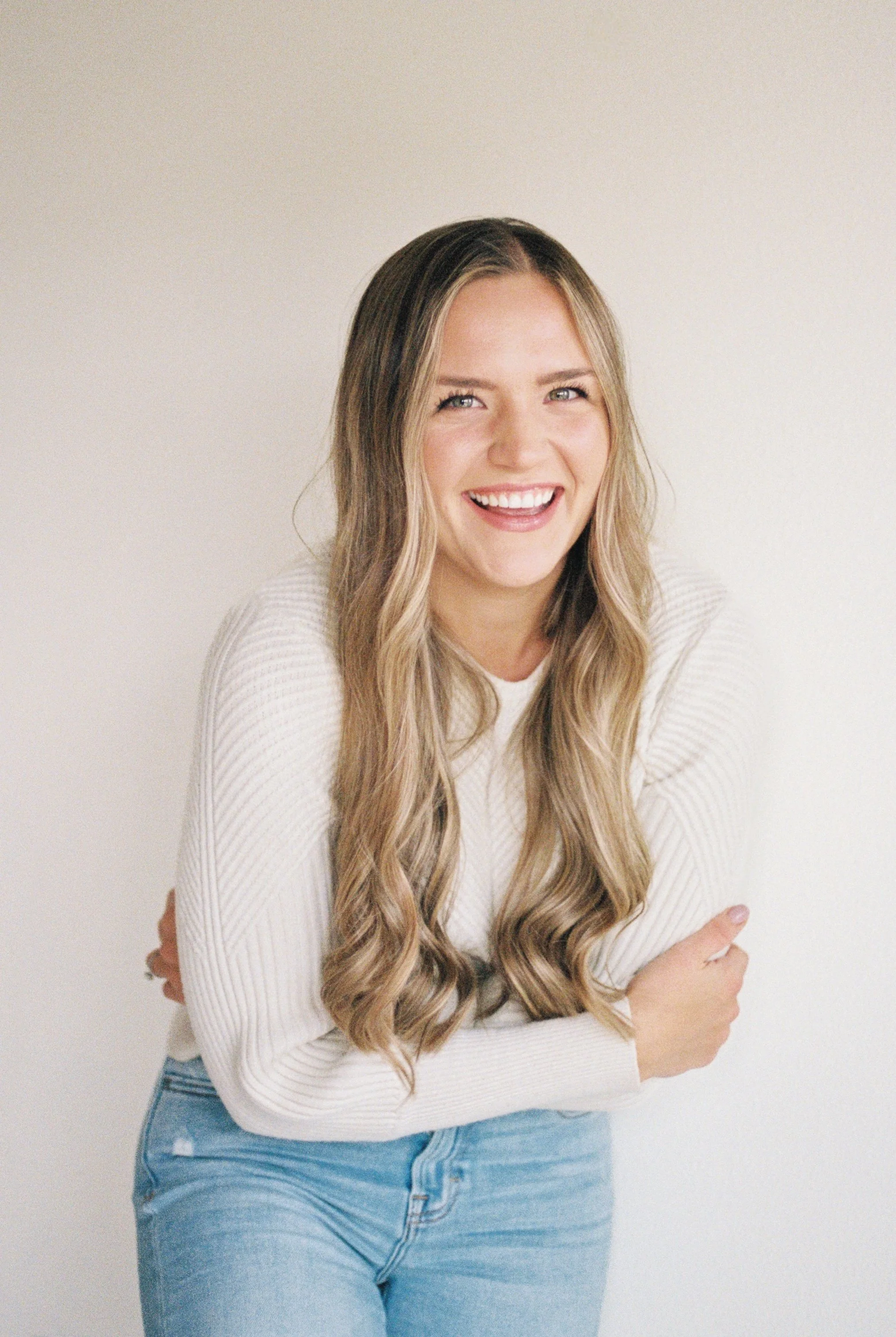A woman with long, wavy blonde hair smiling and crossing her arms, wearing a white sweater and blue jeans against a plain white background.