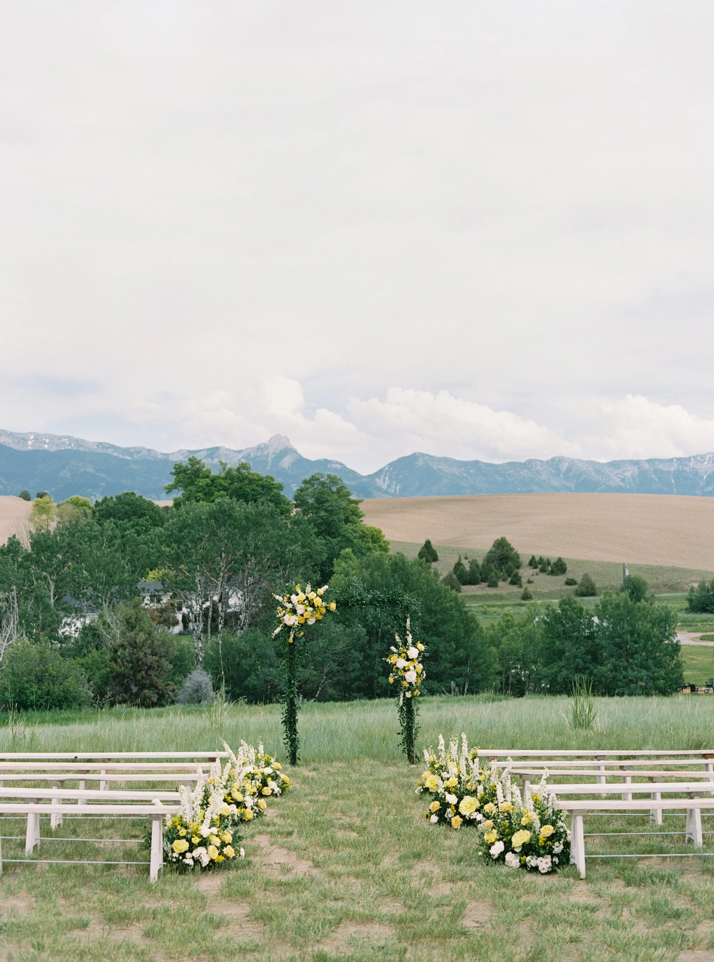 An outdoor wedding setup with white benches on either side of a grassy aisle. The aisle is decorated with bouquets of white and yellow flowers and framed by an arch of greenery and flowers. The scene is set against a backdrop of trees, hills, and distant mountains under a cloudy sky.