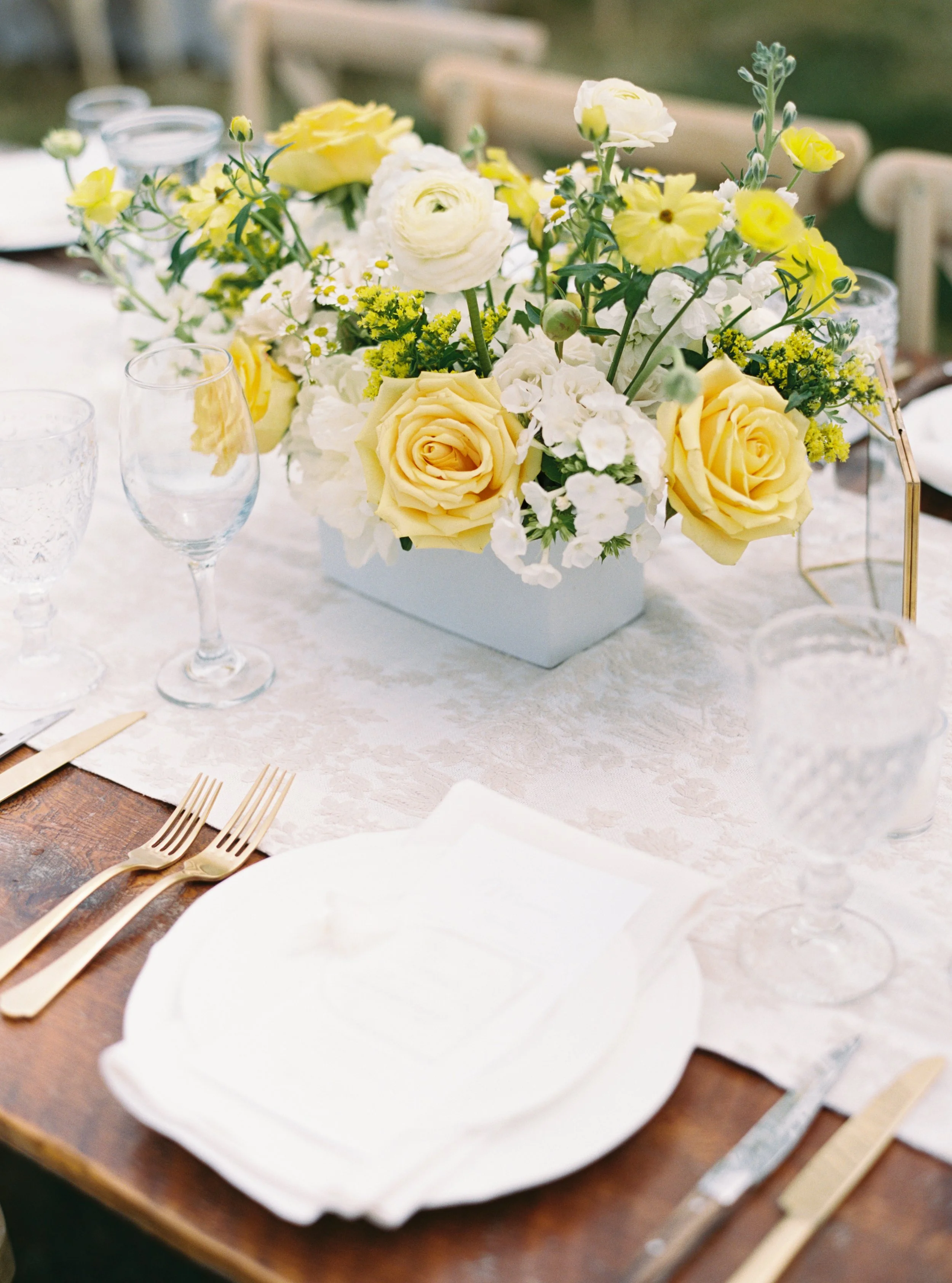 A beautifully set dining table with elegant glassware, gold cutlery, and a floral centerpiece featuring yellow and white roses, complemented by white napkins and plates, suggesting a formal or special occasion.