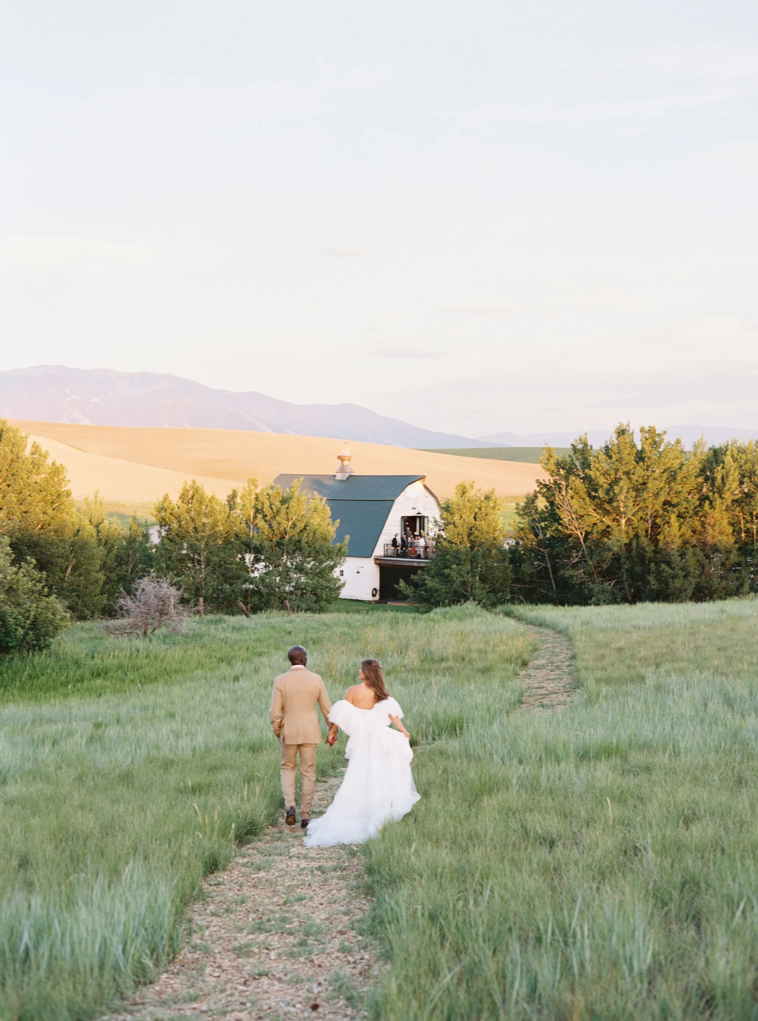 A newly married couple walking hand in hand down a grassy dirt path towards a white barn with a green roof in a rural countryside setting, surrounded by trees and distant mountains in the background.