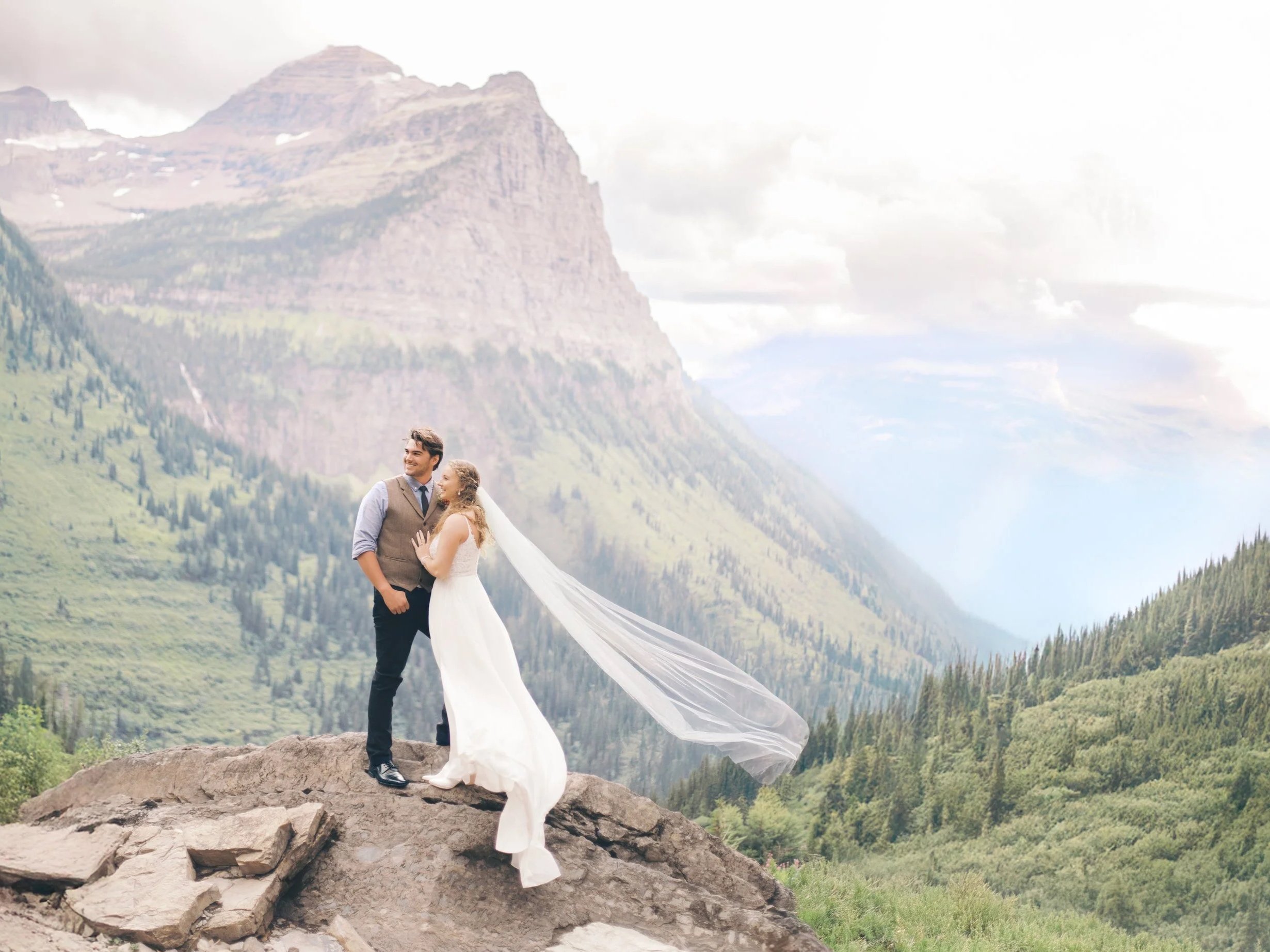 A newlywed couple stands on a rocky ledge in a mountain landscape, with green hills and mountain peaks in the background. The bride wears a white wedding dress with a long veil, and the groom is dressed in a vest and dress shirt, smiling at each other.