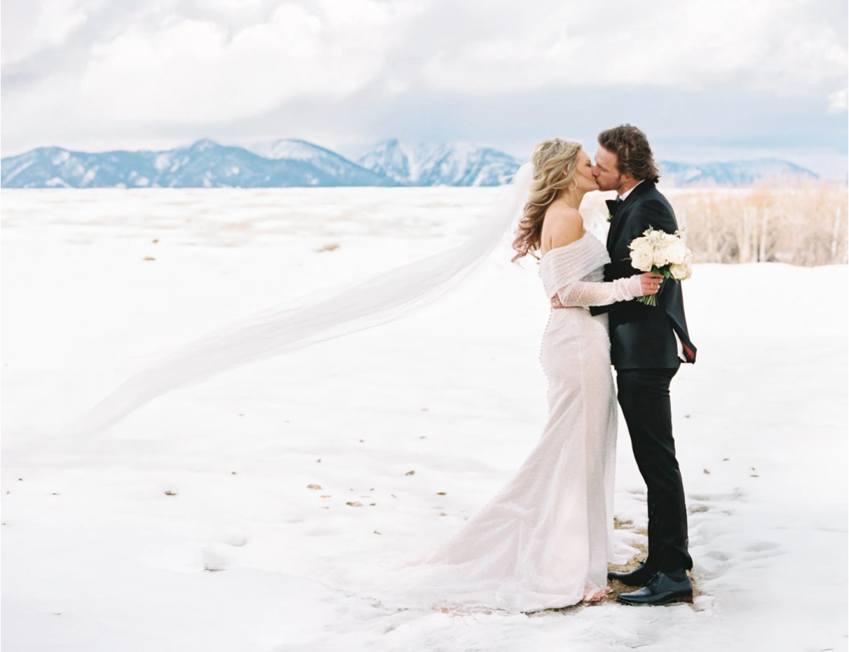 Bride and Groom in Bozeman Montana standing on snow with mountains in background sharing a kiss. Bride is in a white wedding gown and holding a bouquet, while the groom is in a black suit. The background features open fields and a partly cloudy sky.