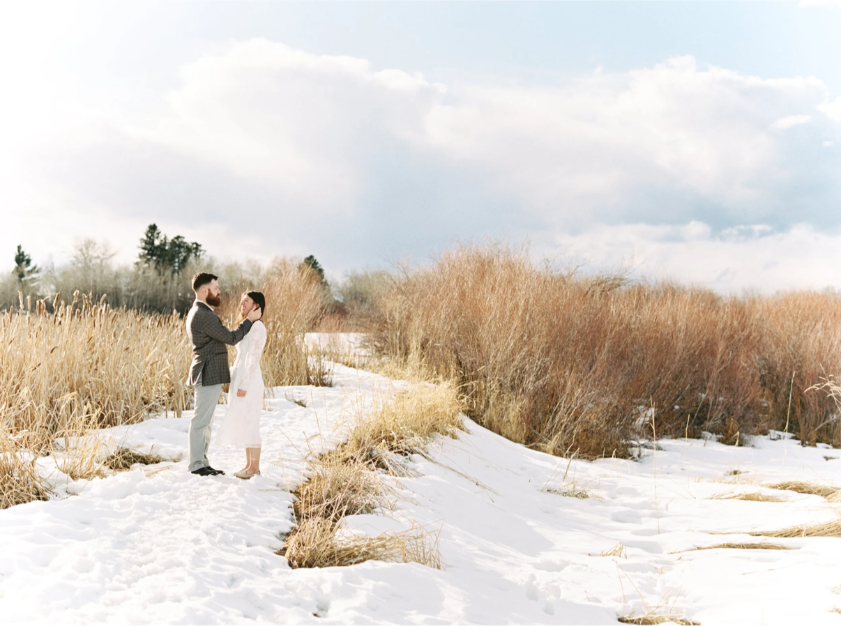 Groom pushing hair behind bride's ear in a snow covered field with tall grass behind them.