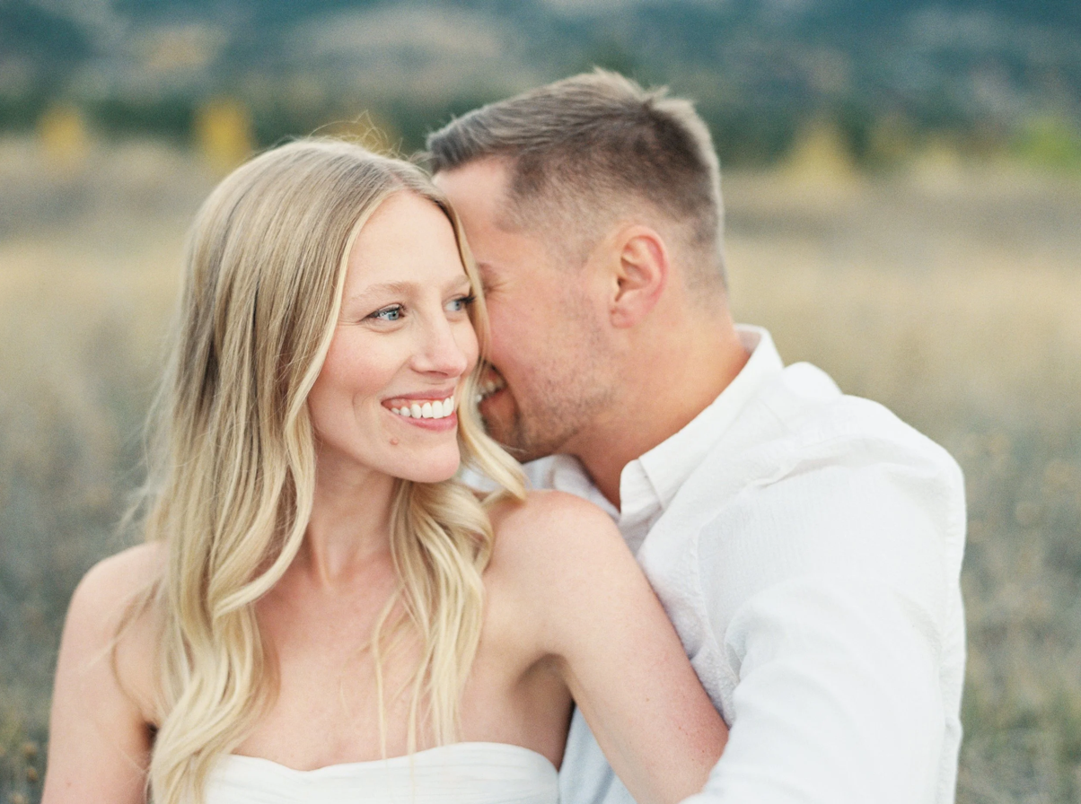 Bride and Groom flirting while sitting in tall grass field with fall yellow trees behind them