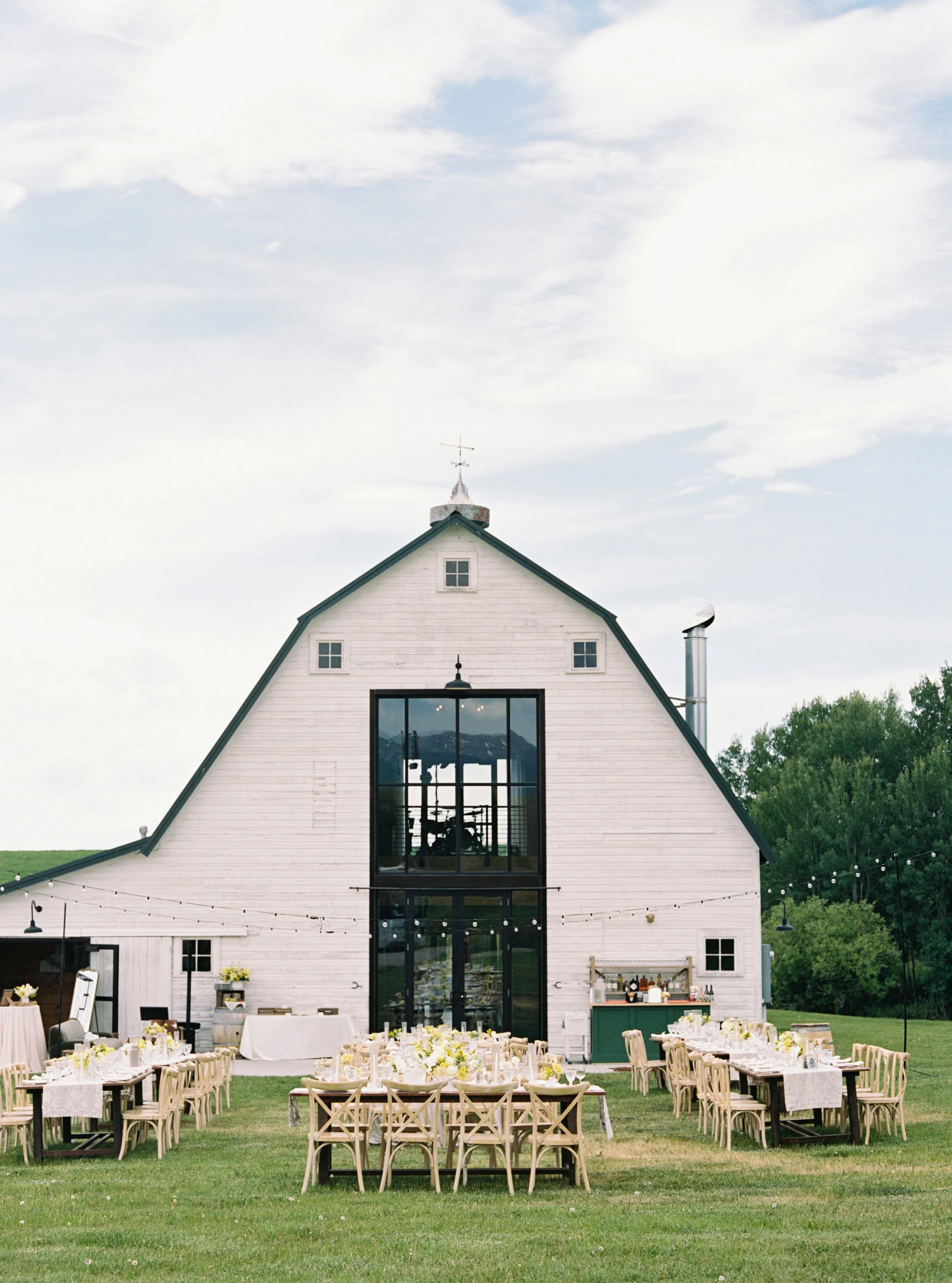 Outdoor wedding reception setup in front of a white barn with tables, chairs, and floral arrangements on a grassy field.